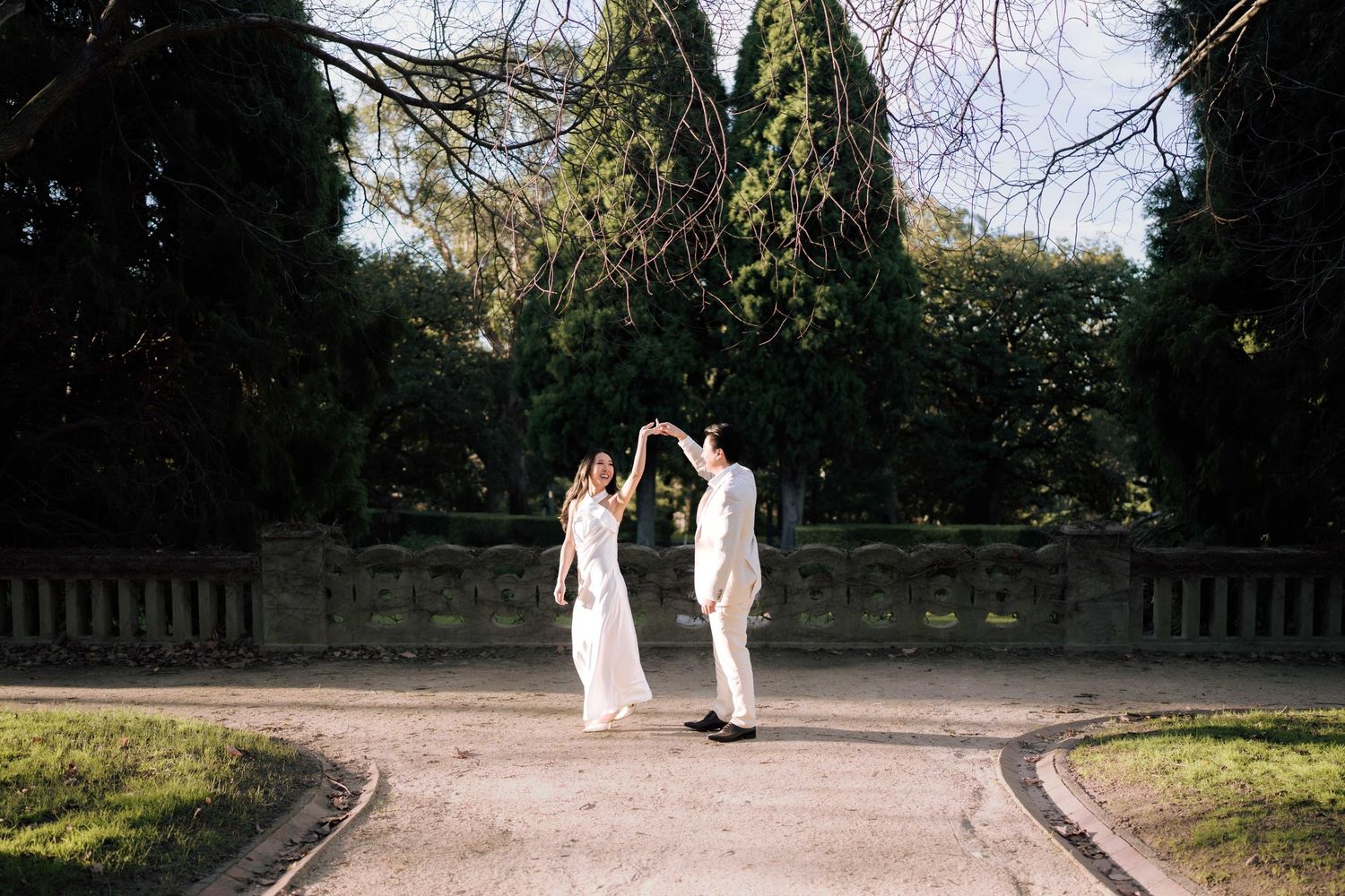 Couple in white wedding attire dancing on garden path surrounded by lush greenery and stone walls.