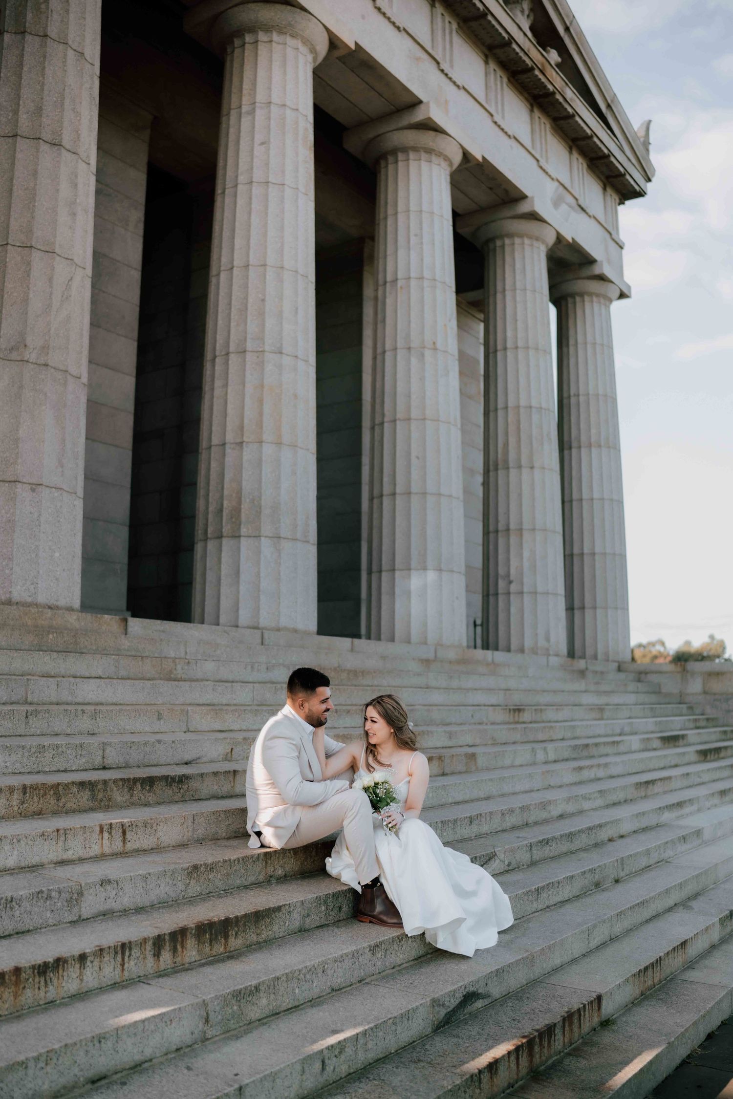 Bride and groom sit on steps of grand columned building sharing an intimate moment on their wedding day.