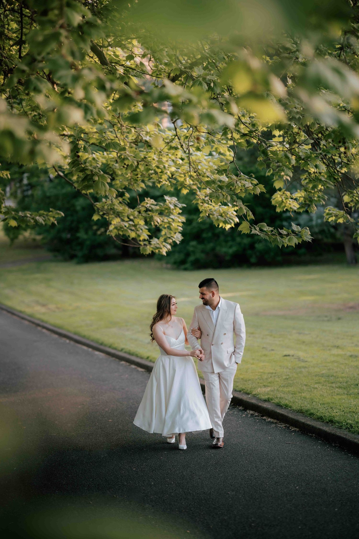 Bride and groom in matching white attire walking hand in hand along tree-lined park pathway.