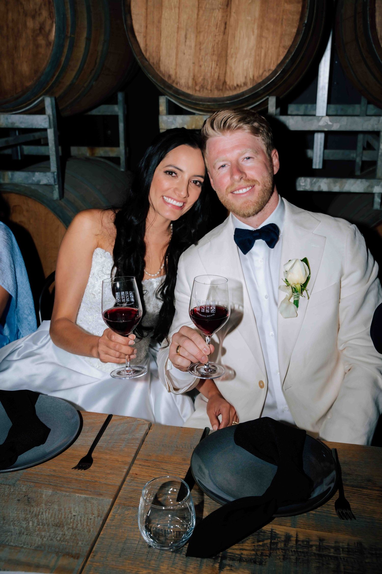Bride and groom toast with red wine glasses seated near oak barrels at winery wedding.