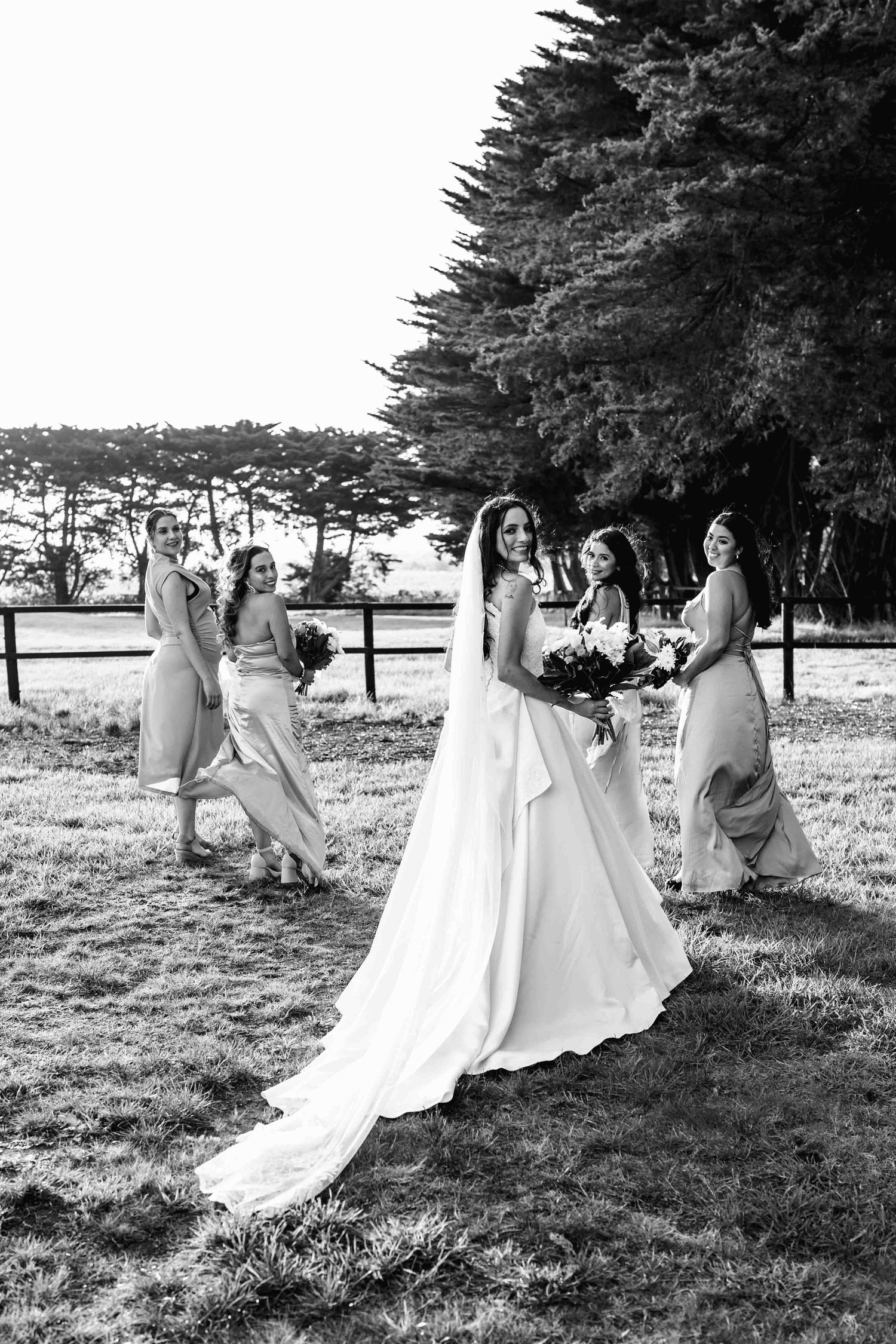 Bride in white gown with long train poses with four bridesmaids in matching dresses outdoors in black and white photo.