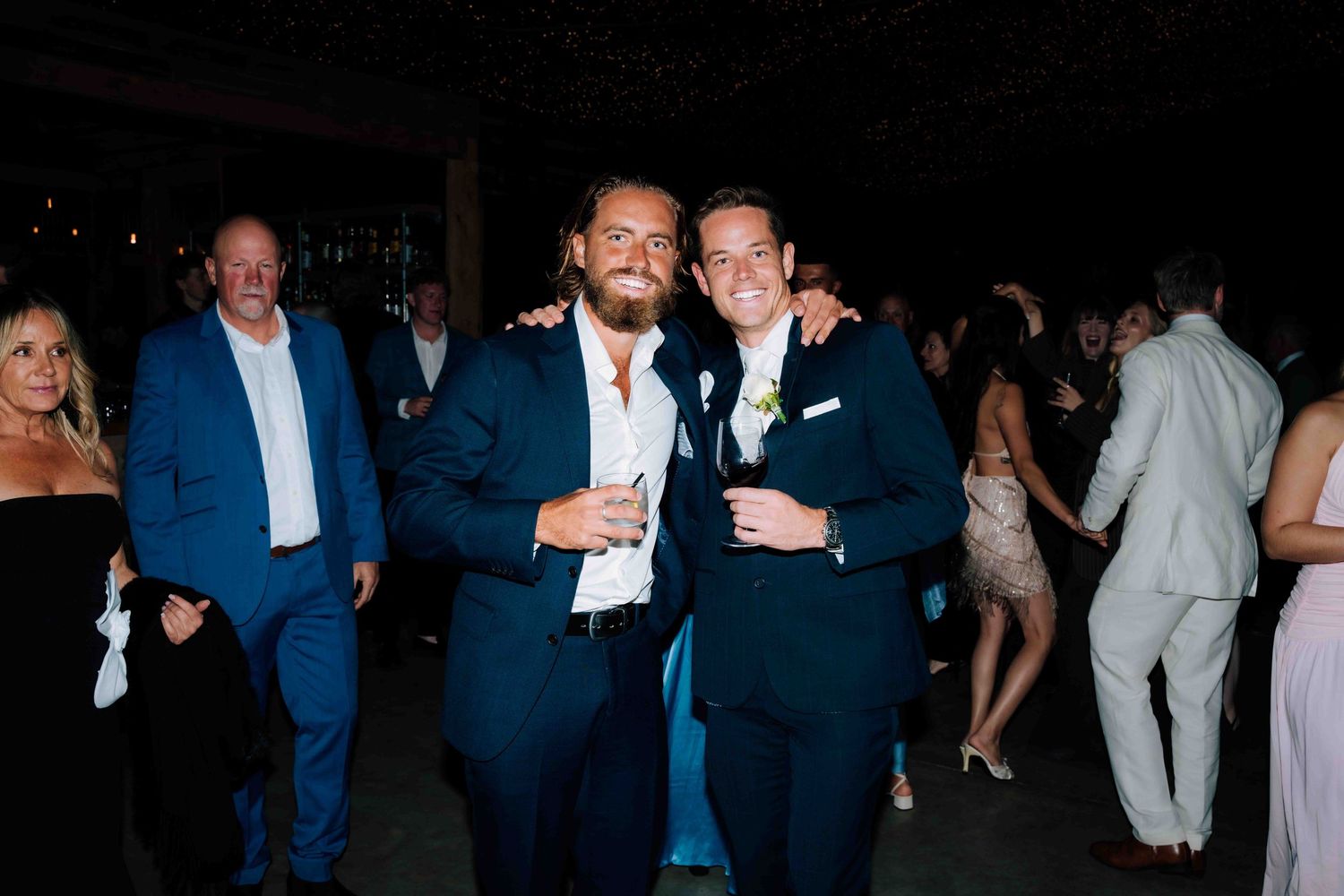 Two men in navy suits smiling and posing together at a wedding reception.