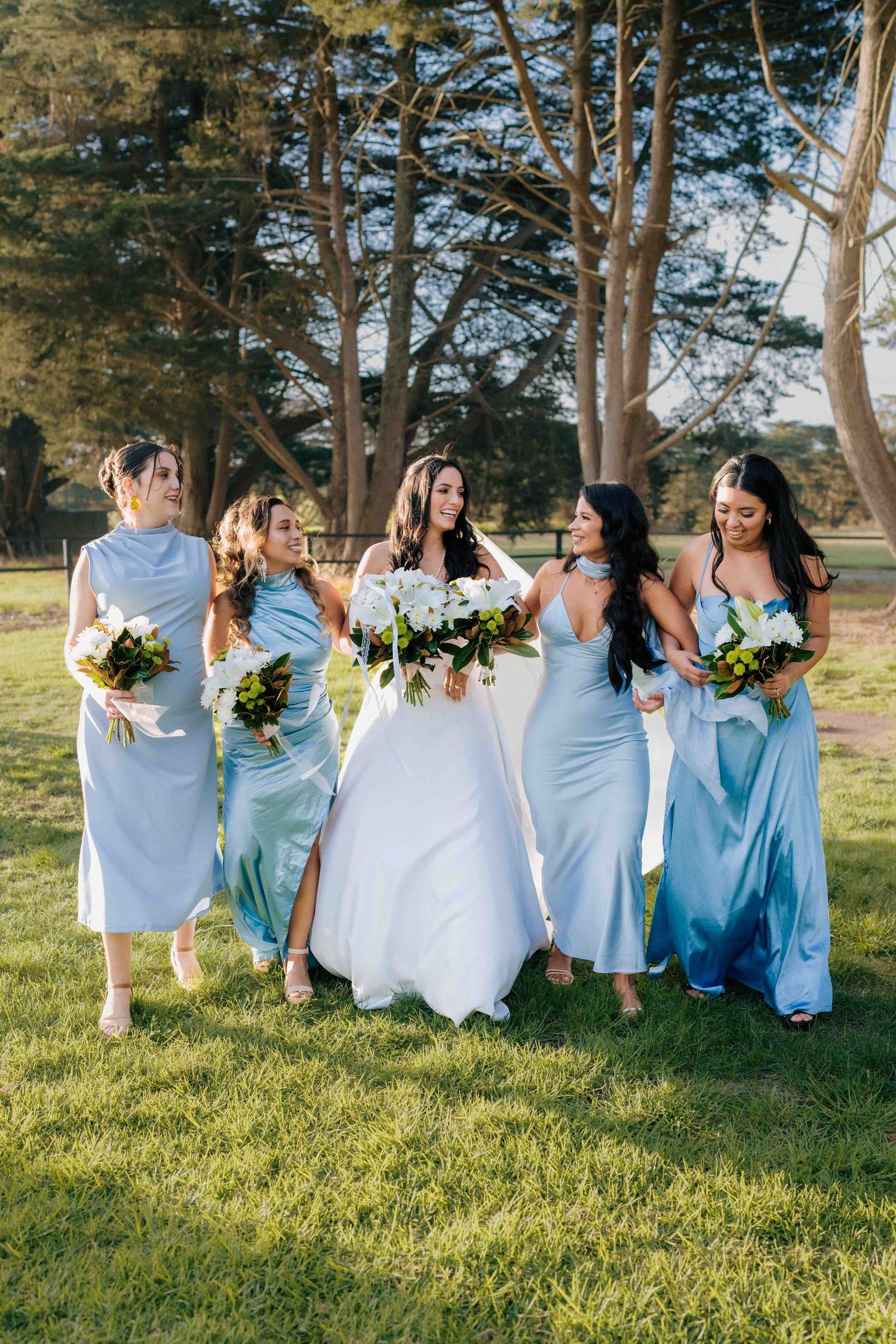 Bride in white gown walking with four bridesmaids in light blue dresses carrying bouquets outdoors.