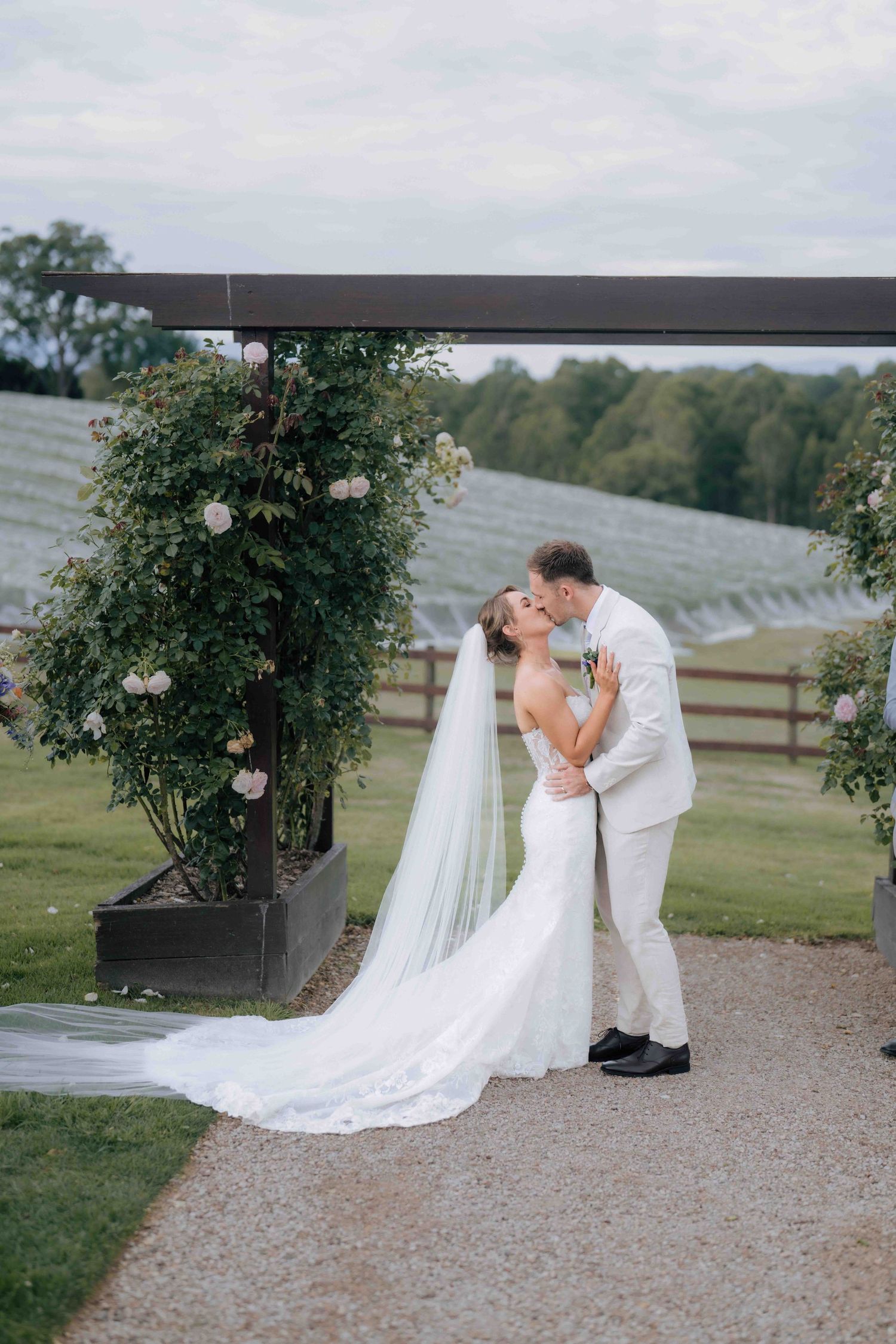 Bride and groom share a romantic kiss under a floral arch at a vineyard wedding ceremony.