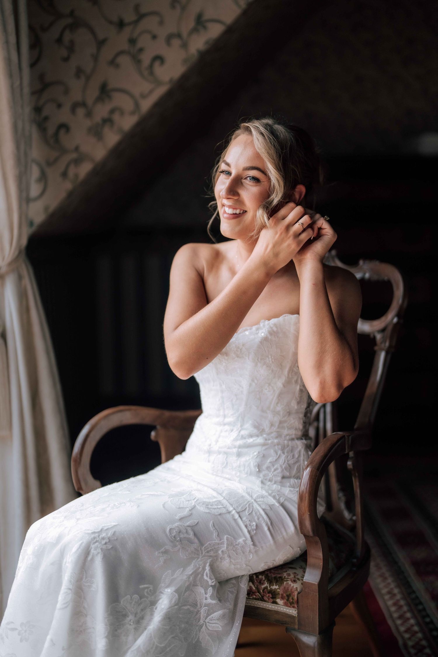 Smiling bride in strapless white lace gown sitting in vintage wooden chair adjusting earring.