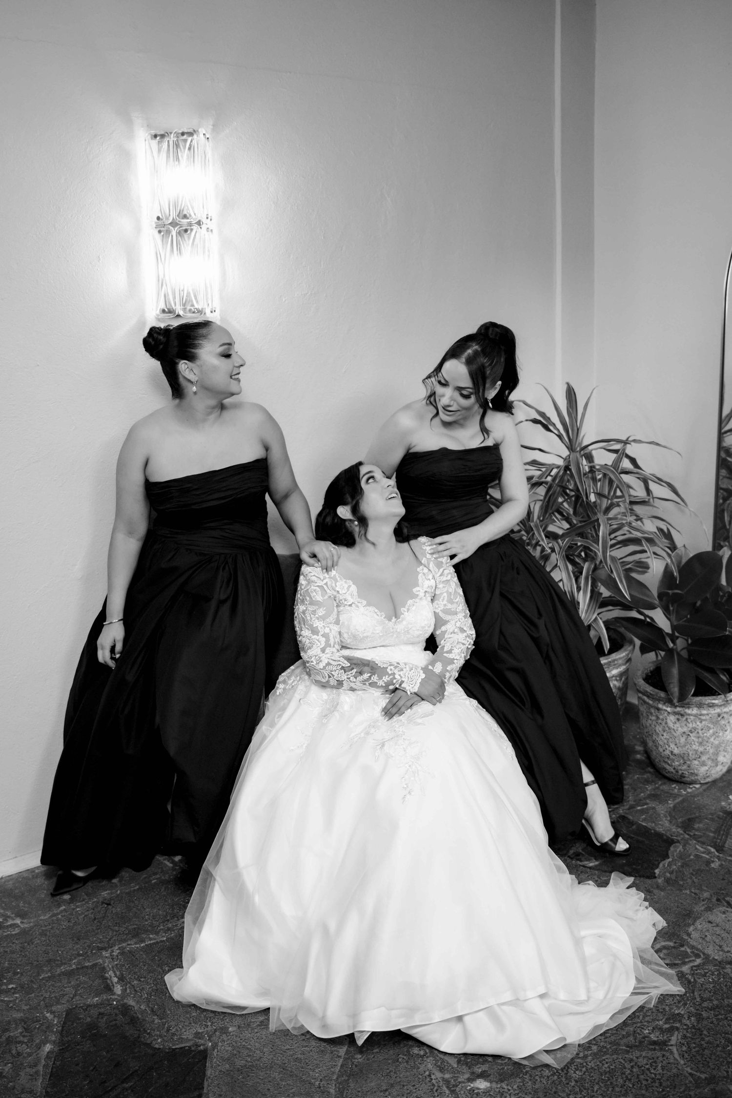 Black and white photo of bride in lace gown seated between two bridesmaids in black dresses.
