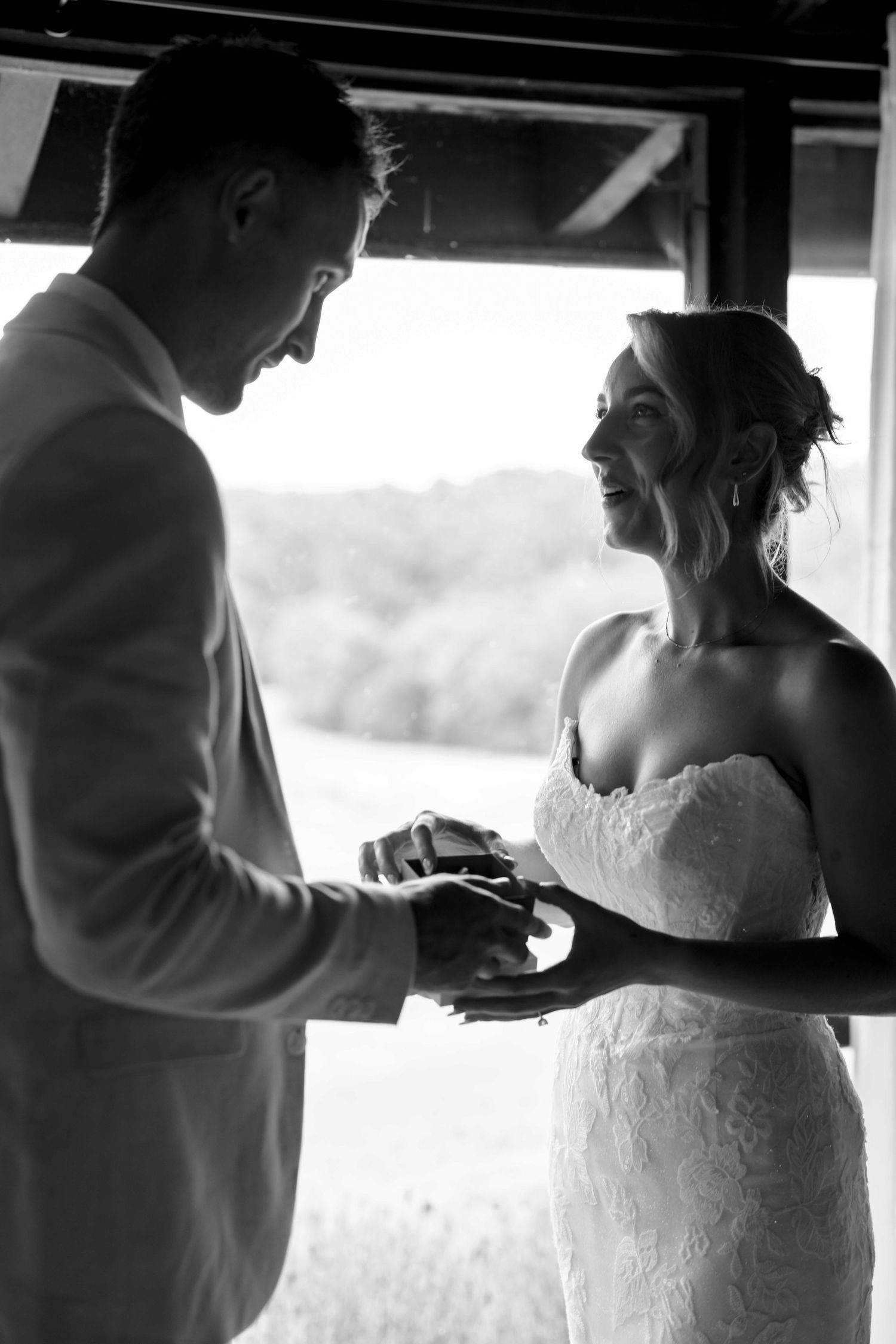 Black and white photo of smiling bride in strapless lace gown receiving a gift box from groom in suit.