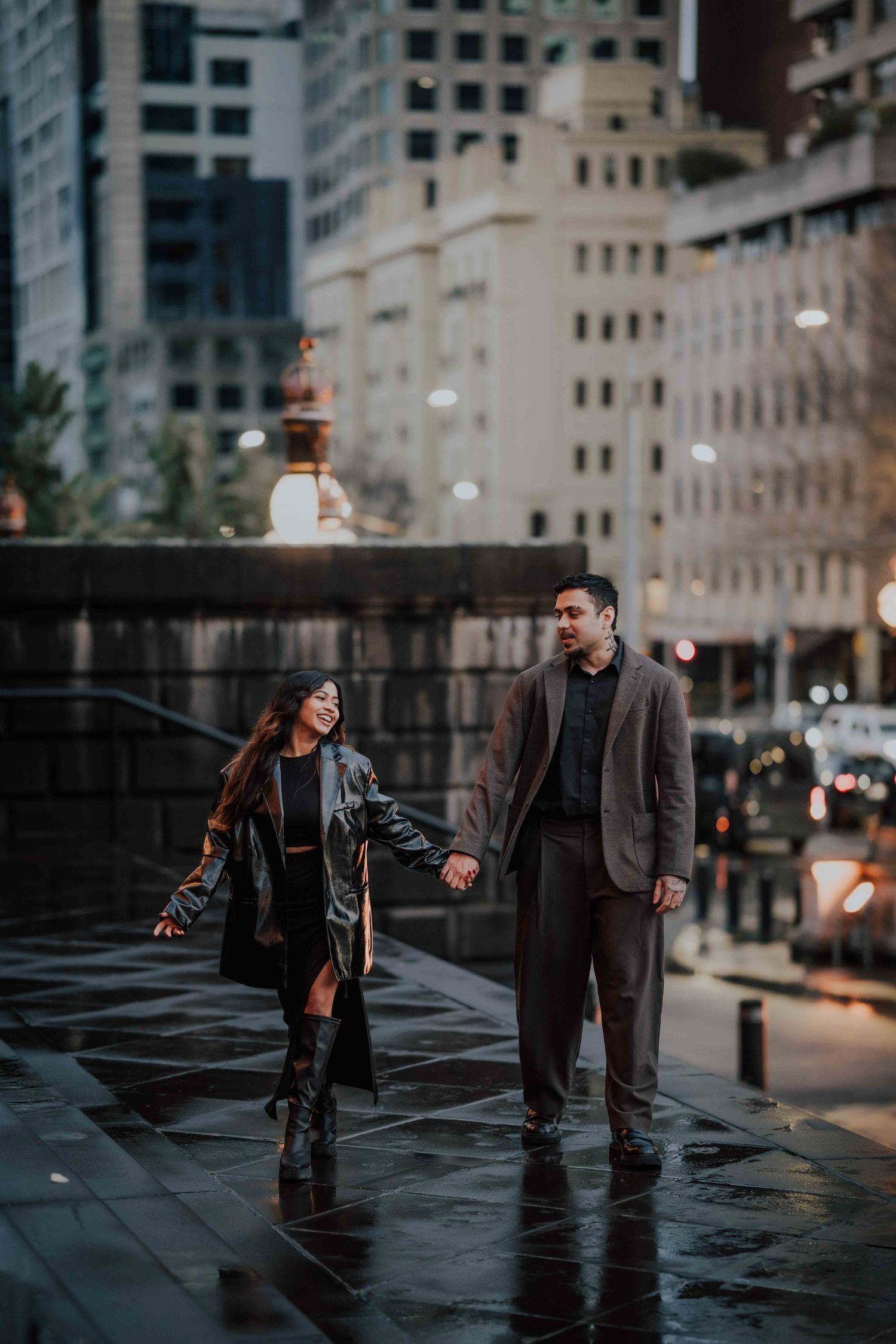 Couple holding hands walking on wet city streets at dusk in stylish outfits.