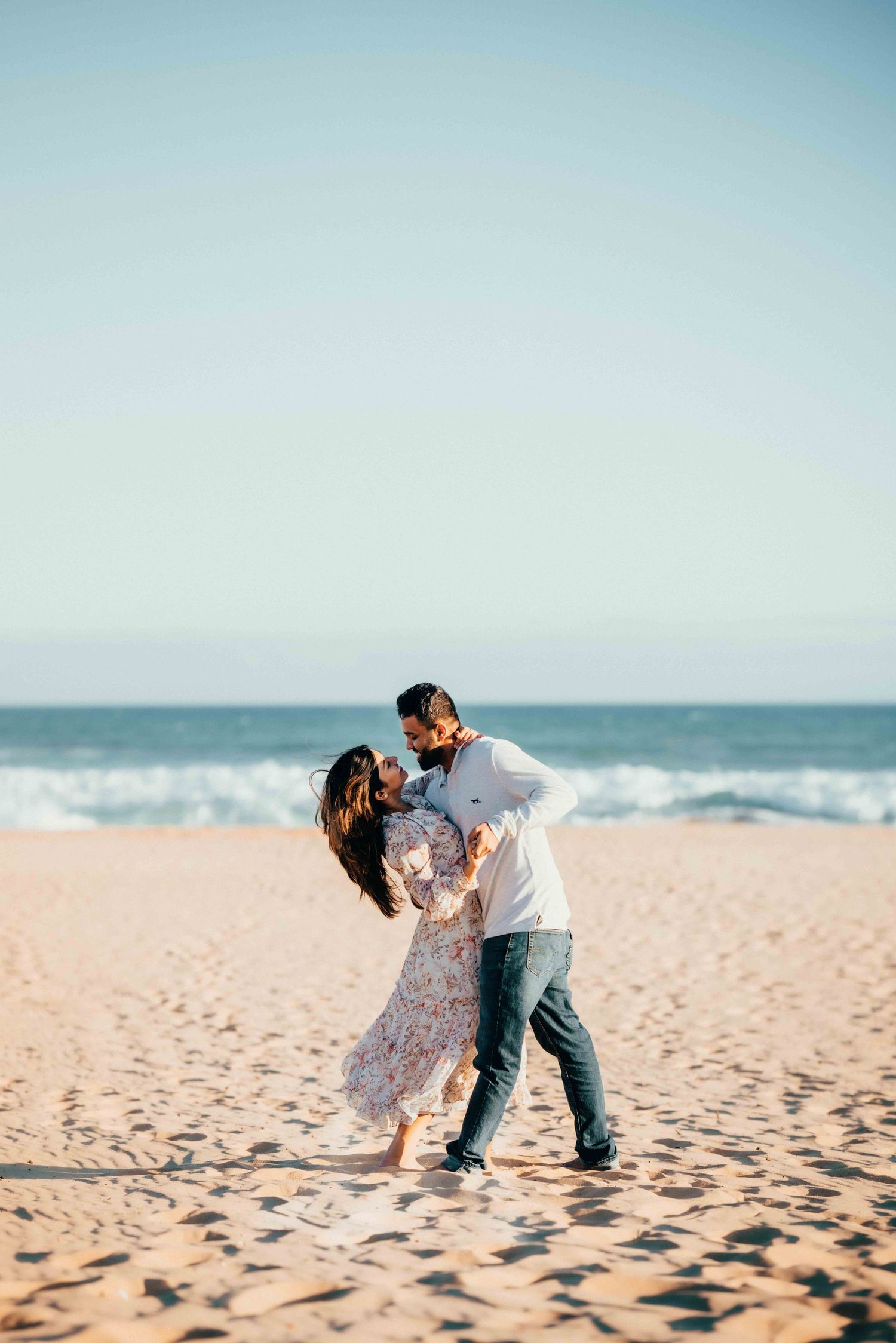 Couple sharing a romantic dip kiss on sunny beach with blue ocean waves and clear sky behind them.