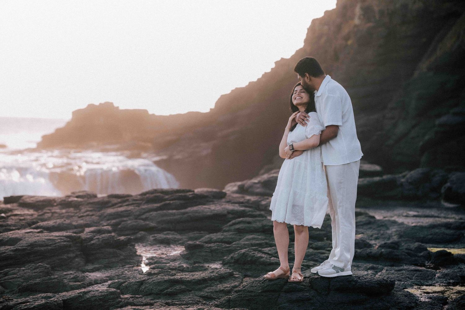 Romantic couple embracing on rocky coast during golden sunset with ocean backdrop.