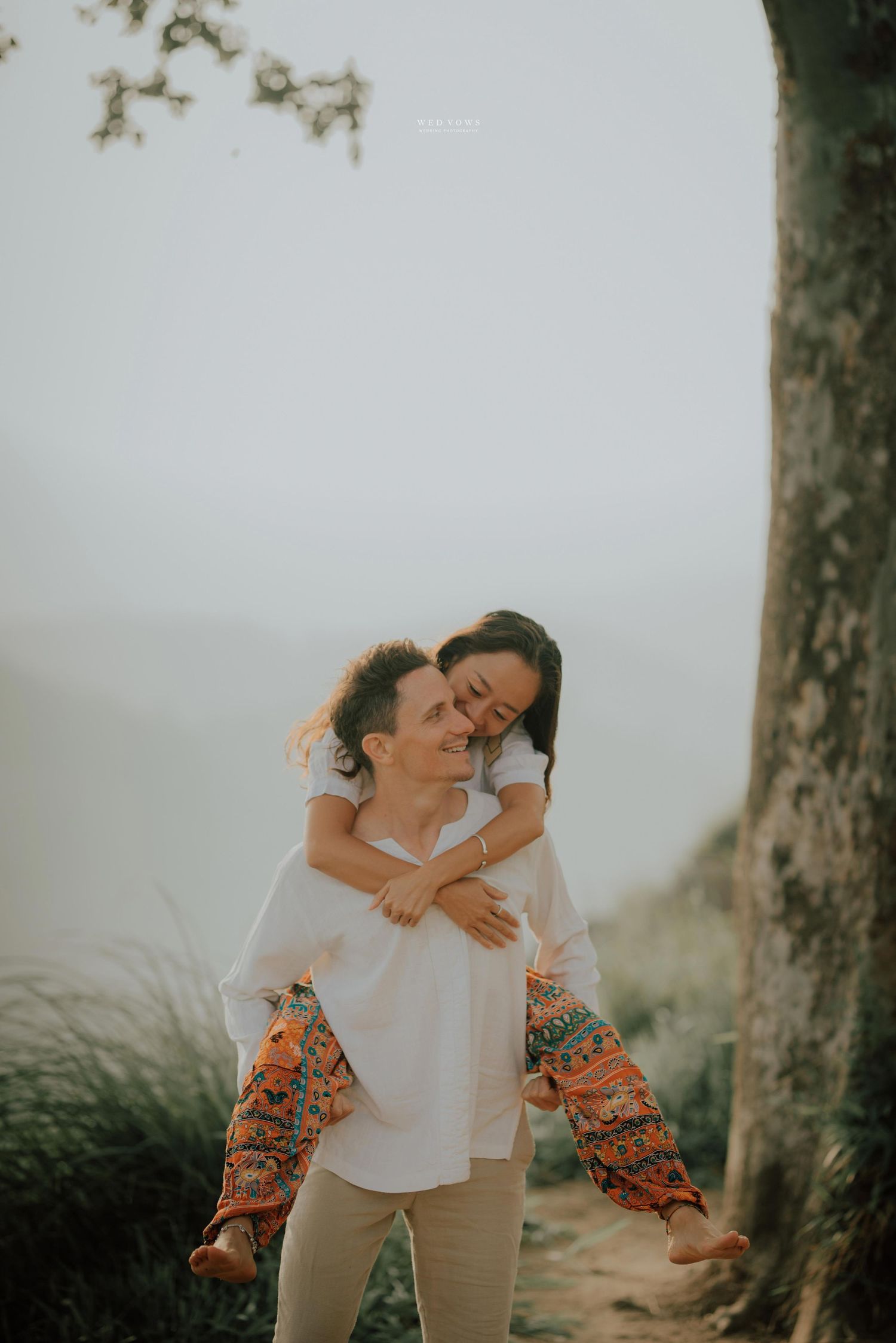 A woman piggybacks on a man in white shirt outdoors with misty background.