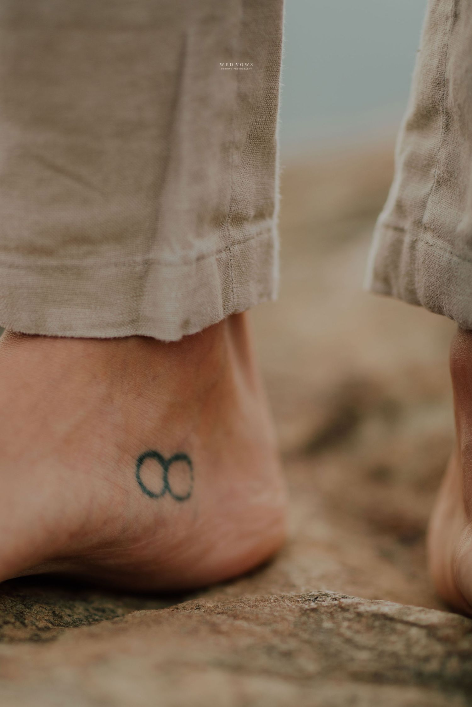Close-up of an ankle tattoo featuring an infinity symbol on a bare foot, wearing beige linen pants on rocky ground.