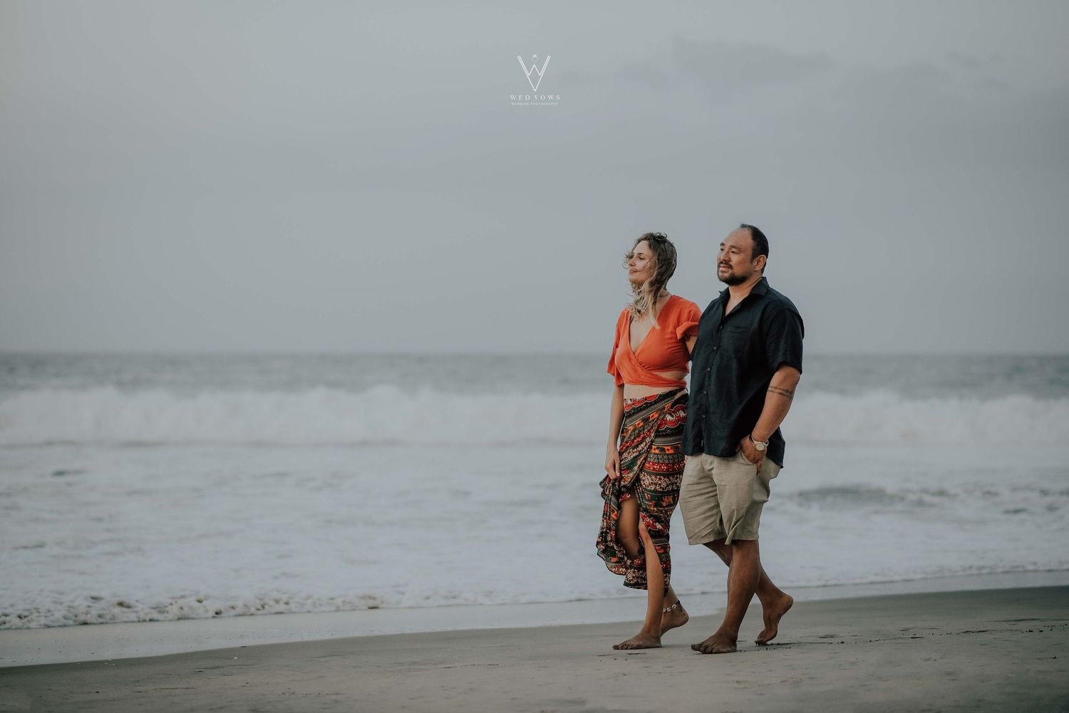 Couple walking hand in hand along a sandy beach with ocean waves in the background.