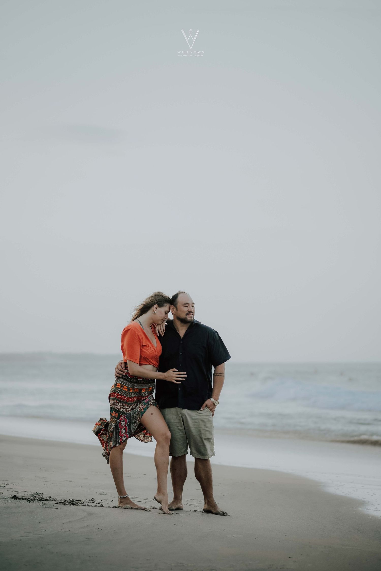 Couple embracing and smiling while walking along a sandy beach with ocean waves.