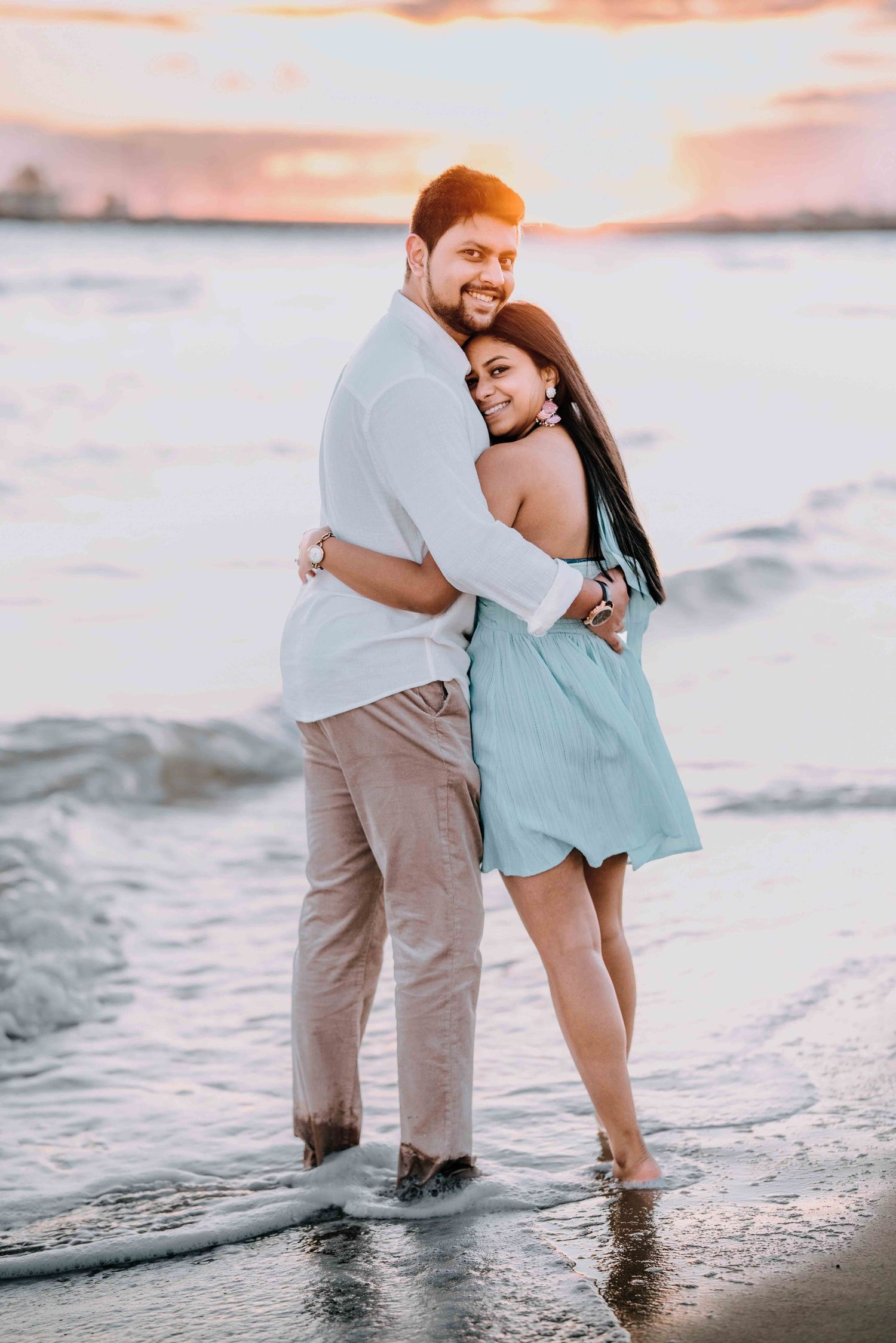 Smiling couple hugging on beach at sunset, woman in light blue dress, golden sky in background.