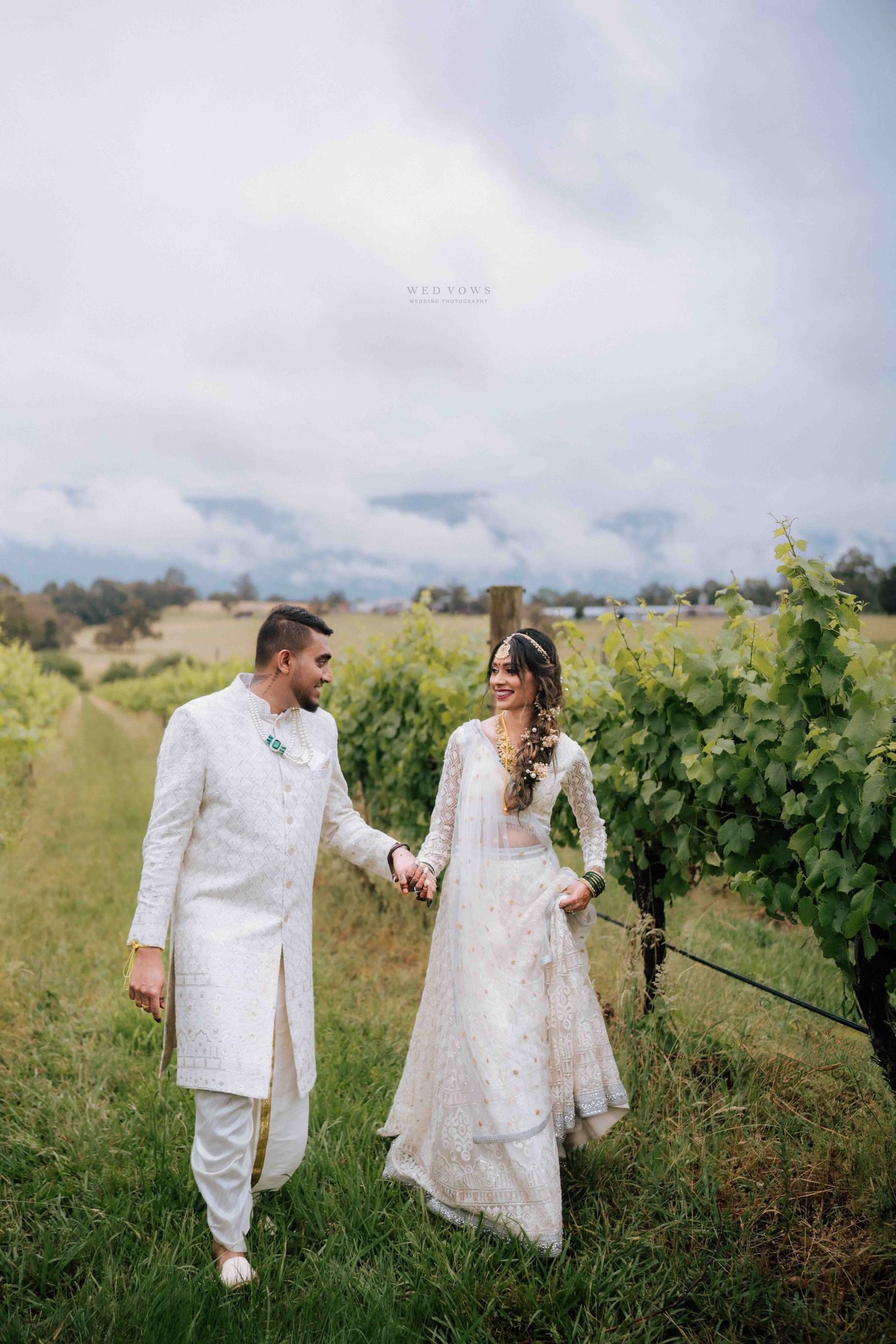 Indian wedding couple in white traditional outfits walk hand in hand through a vineyard.