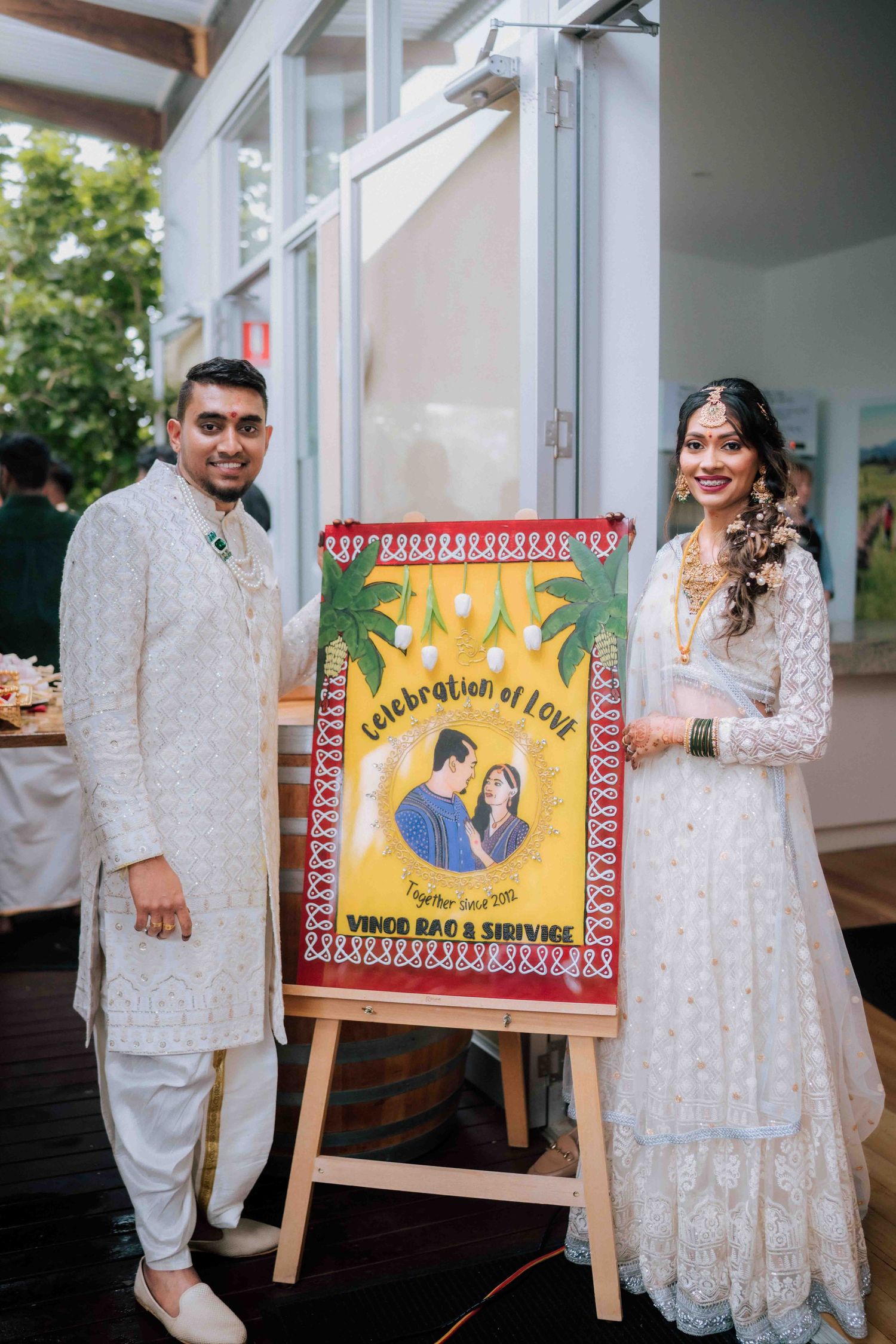 Indian bride and groom posing beside a colorful illustrated wedding sign reading Celebration of Love at their reception.