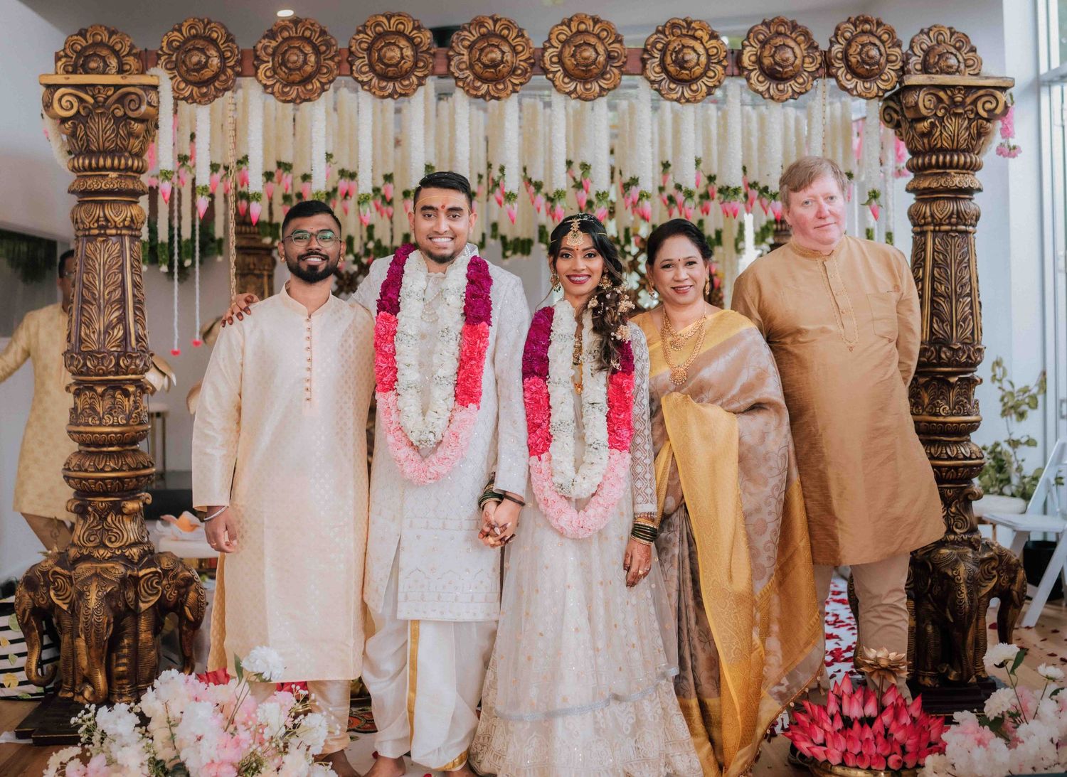 Indian newlyweds with floral garlands pose with four family members in front of an ornate golden wedding mandap.