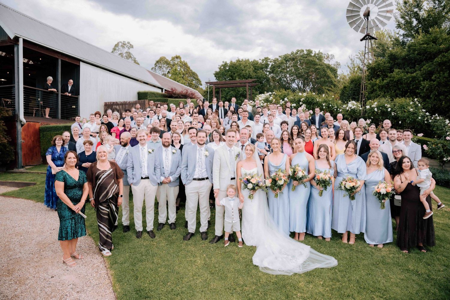 Large wedding group photo with bride, groom, bridesmaids in blue dresses, and guests outside rustic barn venue.