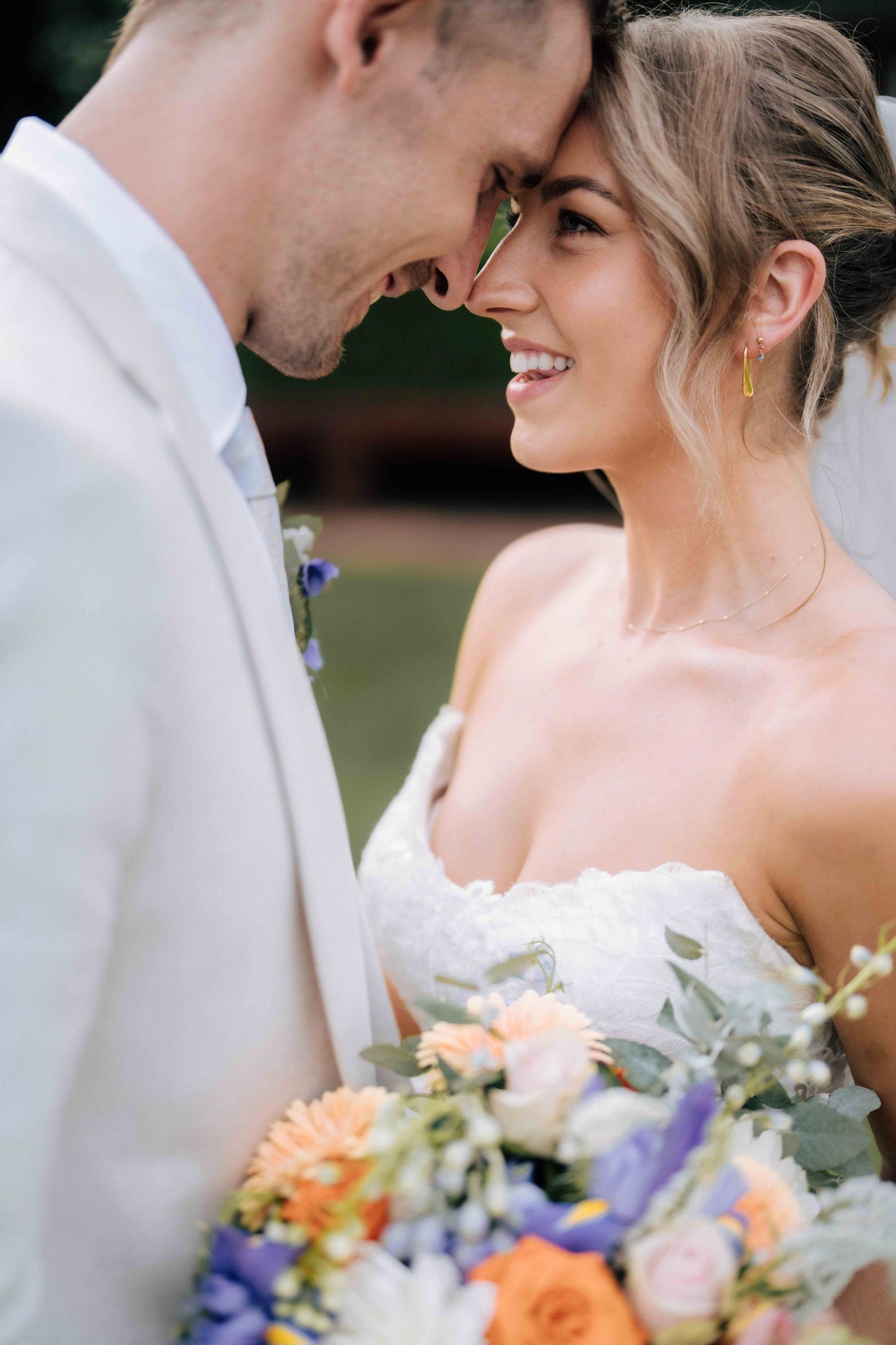 Bride and groom touching foreheads, smiling intimately, bride holding colorful bouquet of orange and purple flowers.