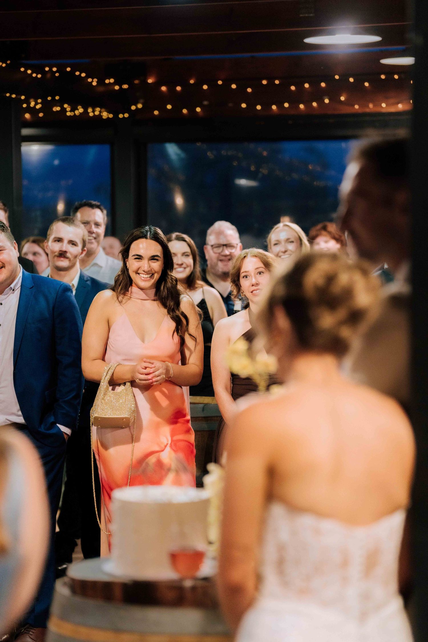 Wedding guest in pink dress smiling at bride during cake cutting ceremony at reception.