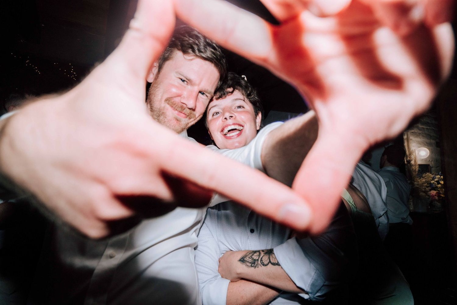 Couple making heart shape with hands toward camera in playful wedding reception photo.