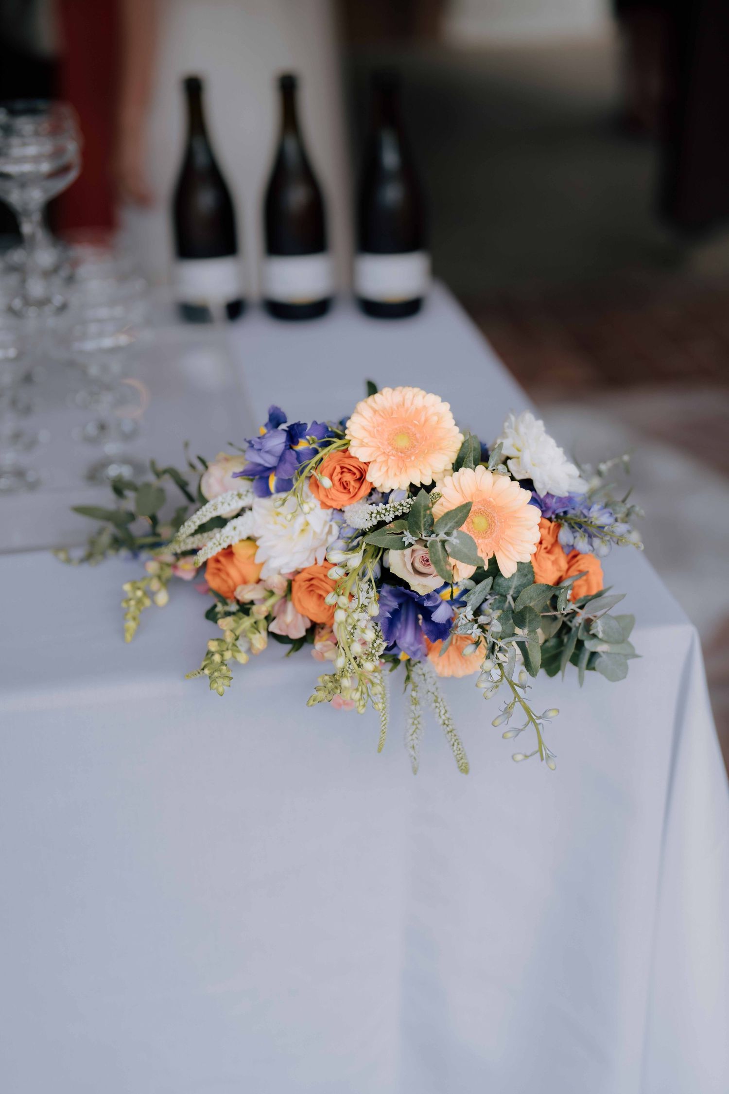 Wedding table centerpiece with orange, purple, and white flowers beside wine bottles and glassware.