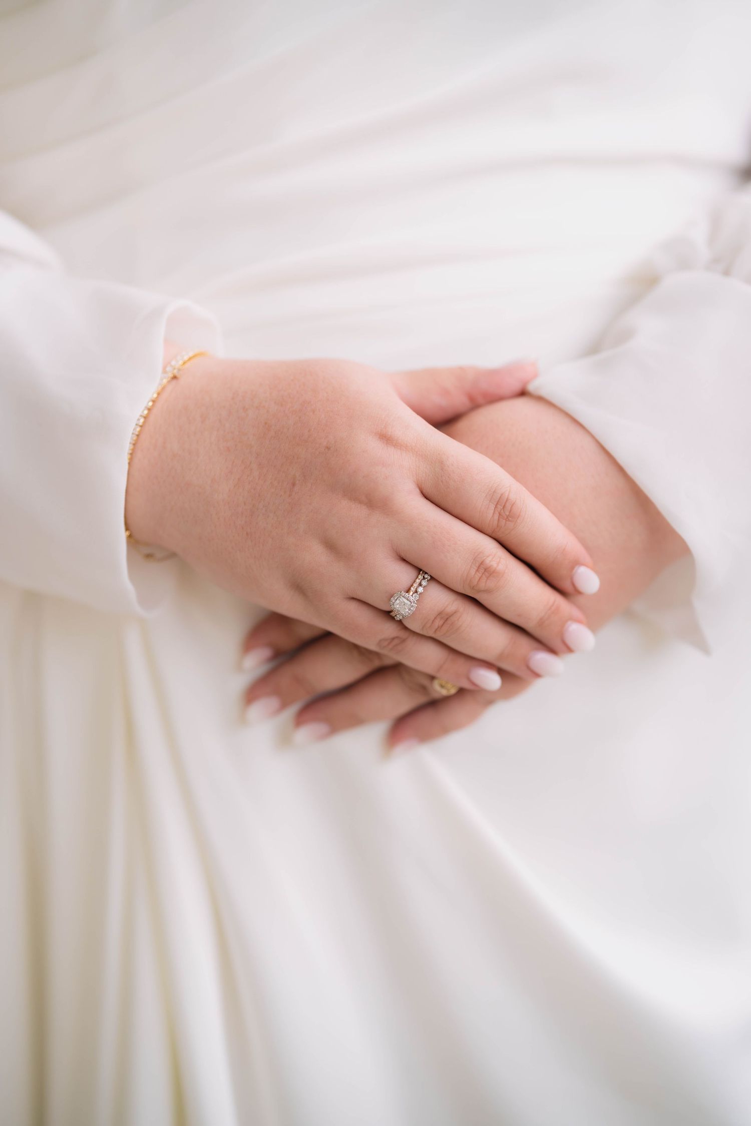 Close-up of bride's hands showing a sparkling diamond engagement ring and gold bracelet on white dress.