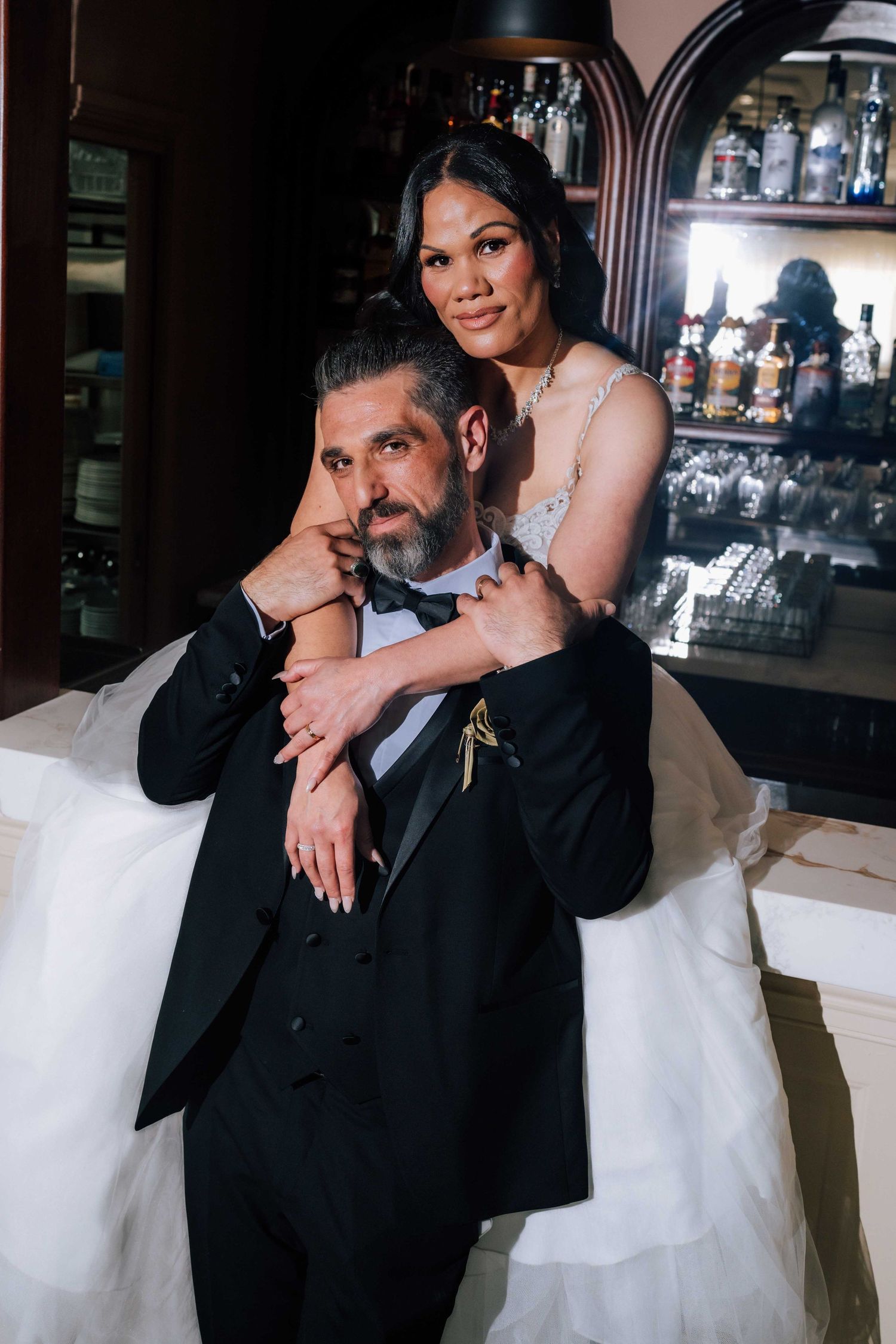 A bride and groom pose intimately together near an elegant bar, with the bride embracing the groom.