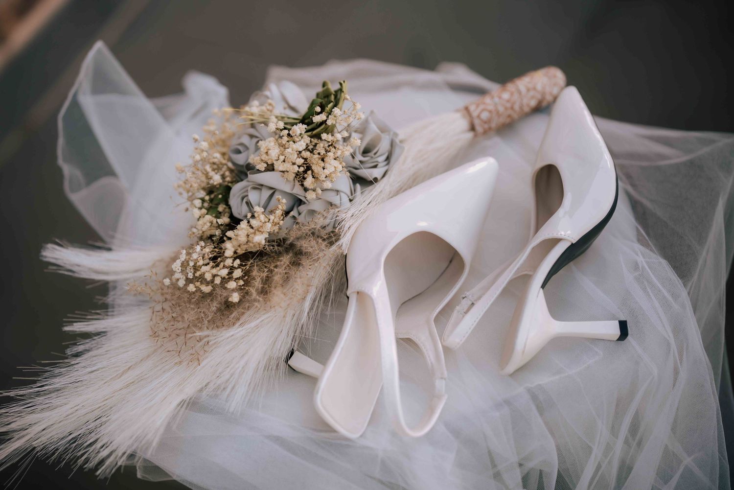 White bridal heels resting on tulle veil beside a dried flower bouquet with feathers.