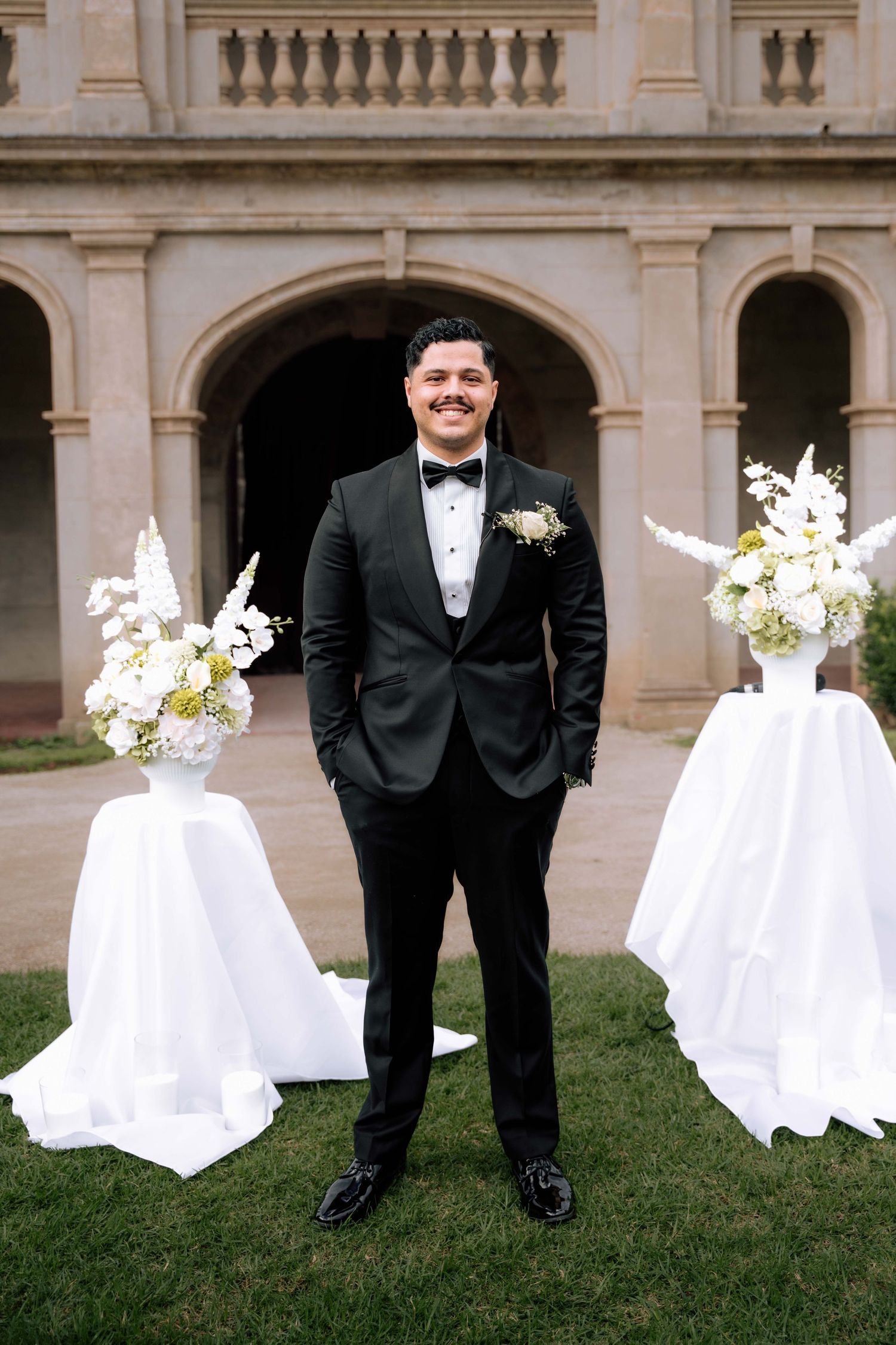 Dapper groom in black tuxedo stands smiling between two white floral arrangements at outdoor ceremony.