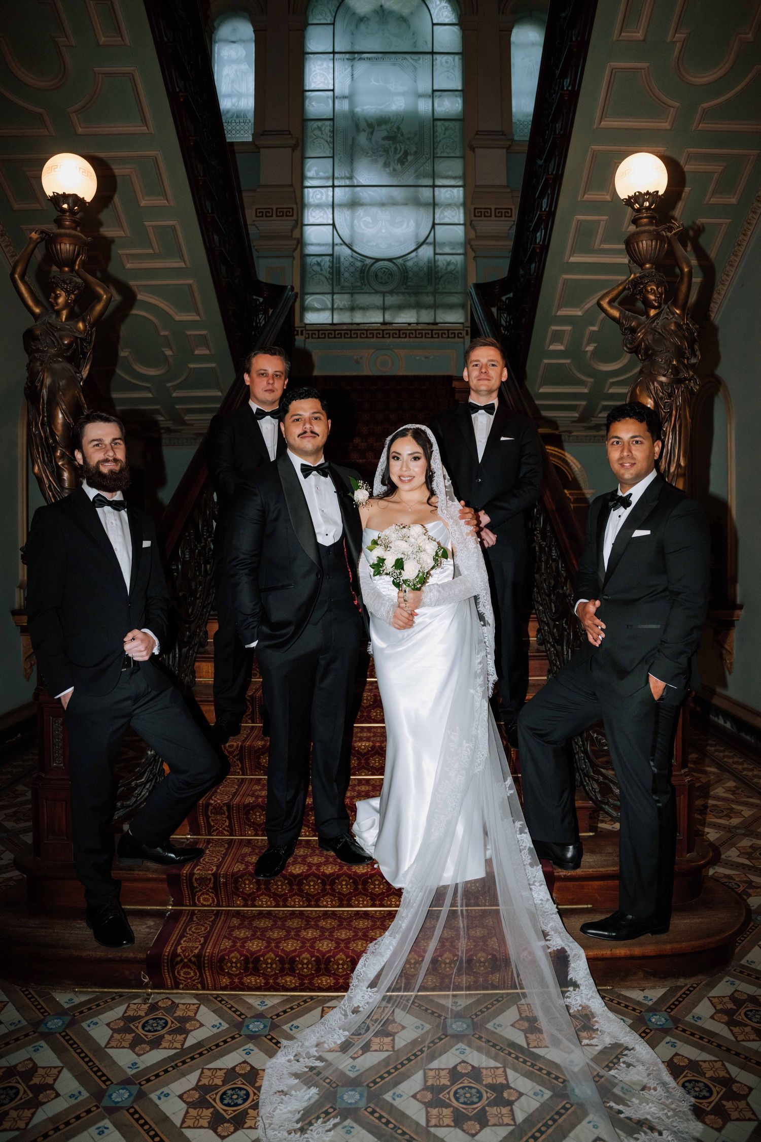 Bride in white gown and veil poses with five groomsmen in black tuxedos on ornate staircase.