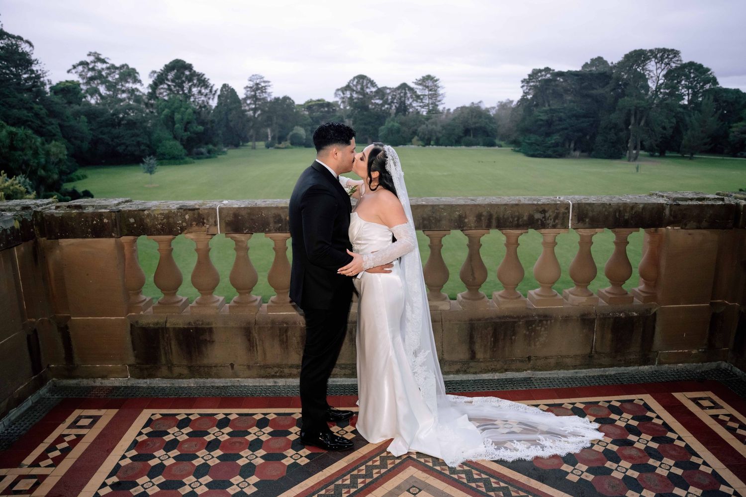 Bride and groom sharing romantic portrait on balcony overlooking lush green garden landscape.