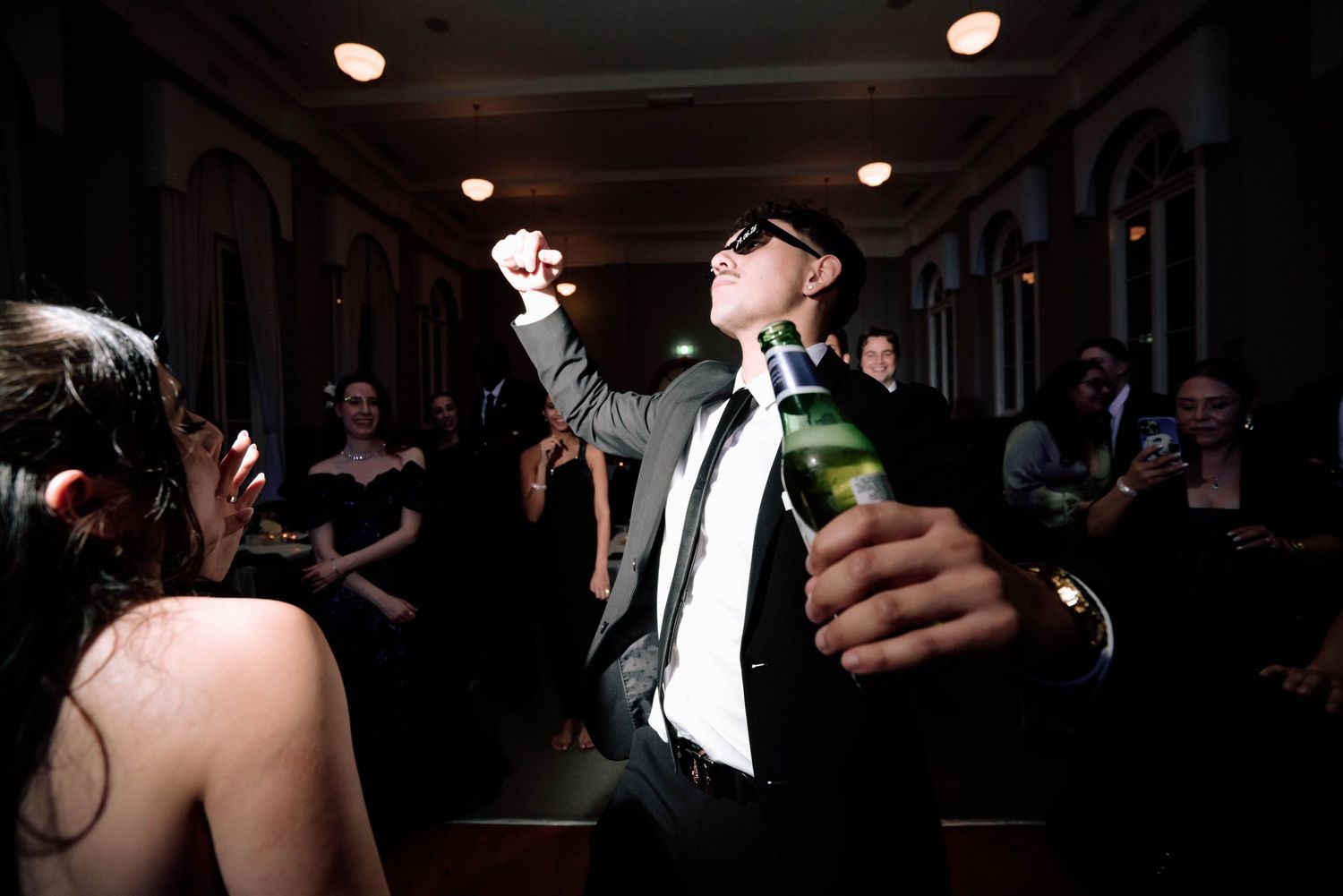 Man in sunglasses and suit dancing energetically while holding a champagne bottle at a party.