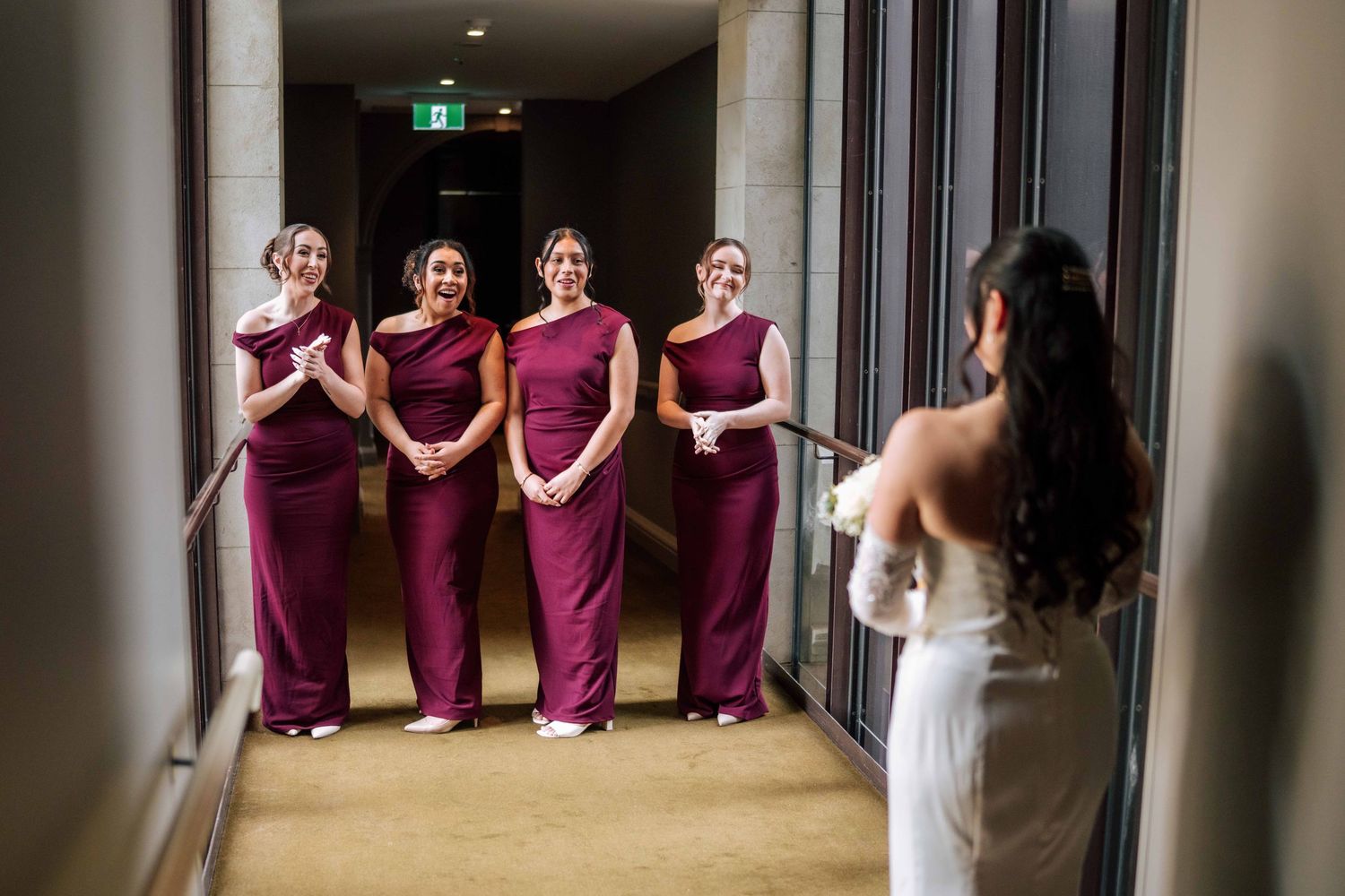 Bride in white gown reveals look to four bridesmaids in burgundy gowns standing in hotel corridor.