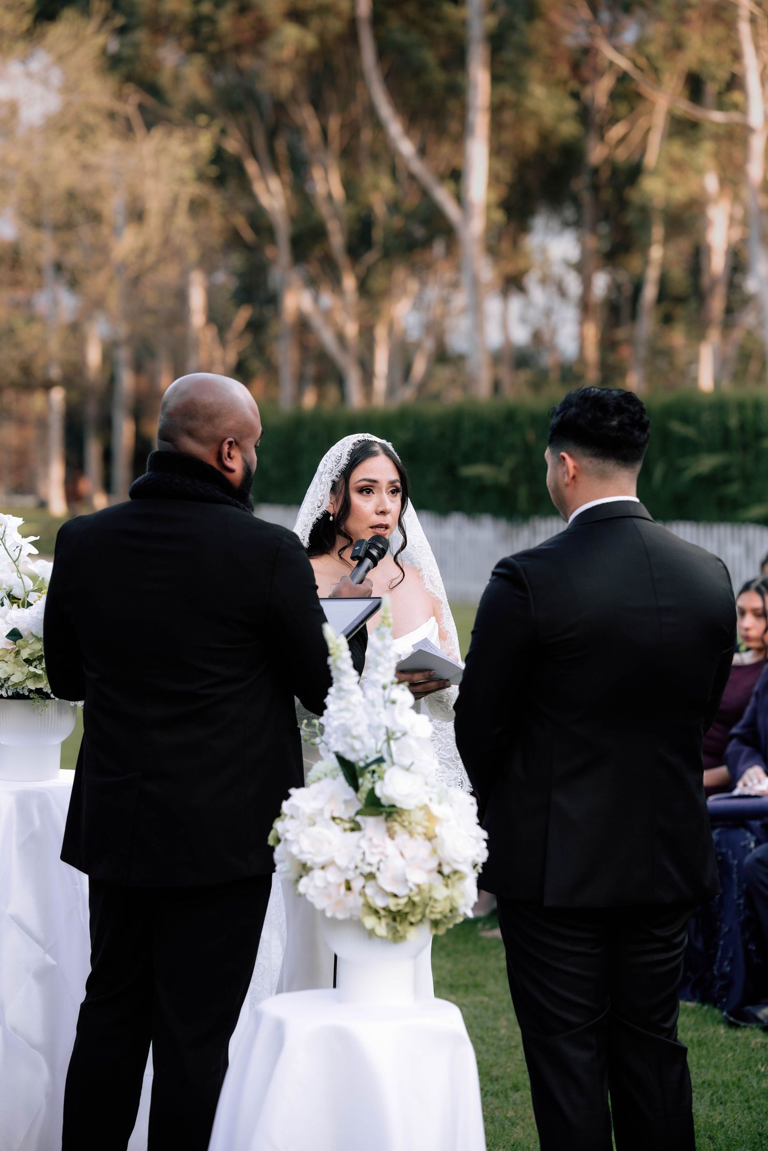 Bride in lace veil reads heartfelt wedding vows to groom at elegant outdoor garden ceremony.