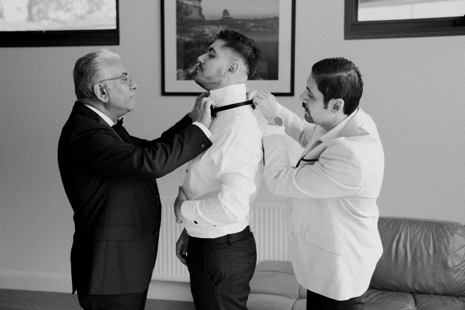 Father and groomsman helping groom fasten bow tie during wedding preparations in black and white.
