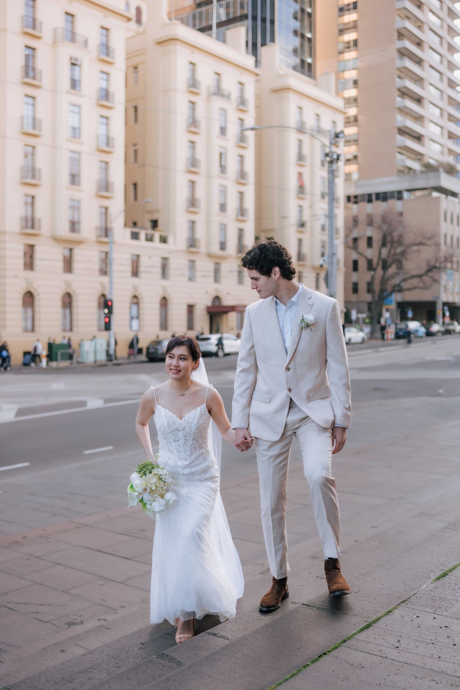 Bride and groom walking hand in hand on city street, bride carrying white bouquet.