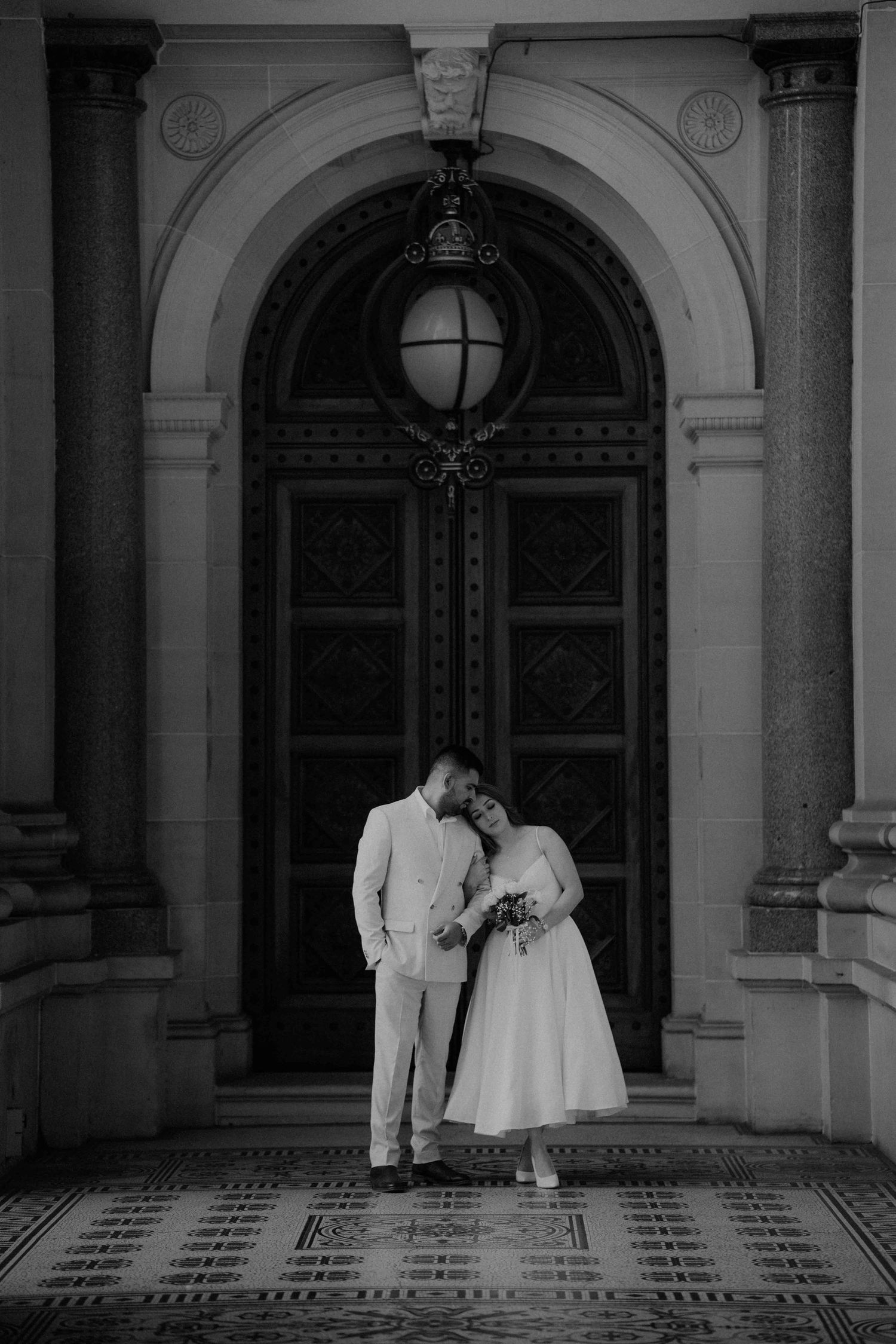 Black and white wedding portrait of a couple embracing before grand ornate doors of a historic building.