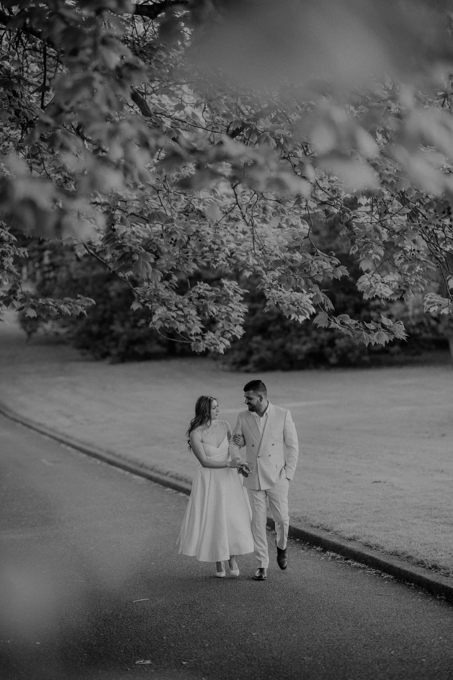 Black and white photo of a wedding couple walking hand in hand along a path surrounded by lush trees.
