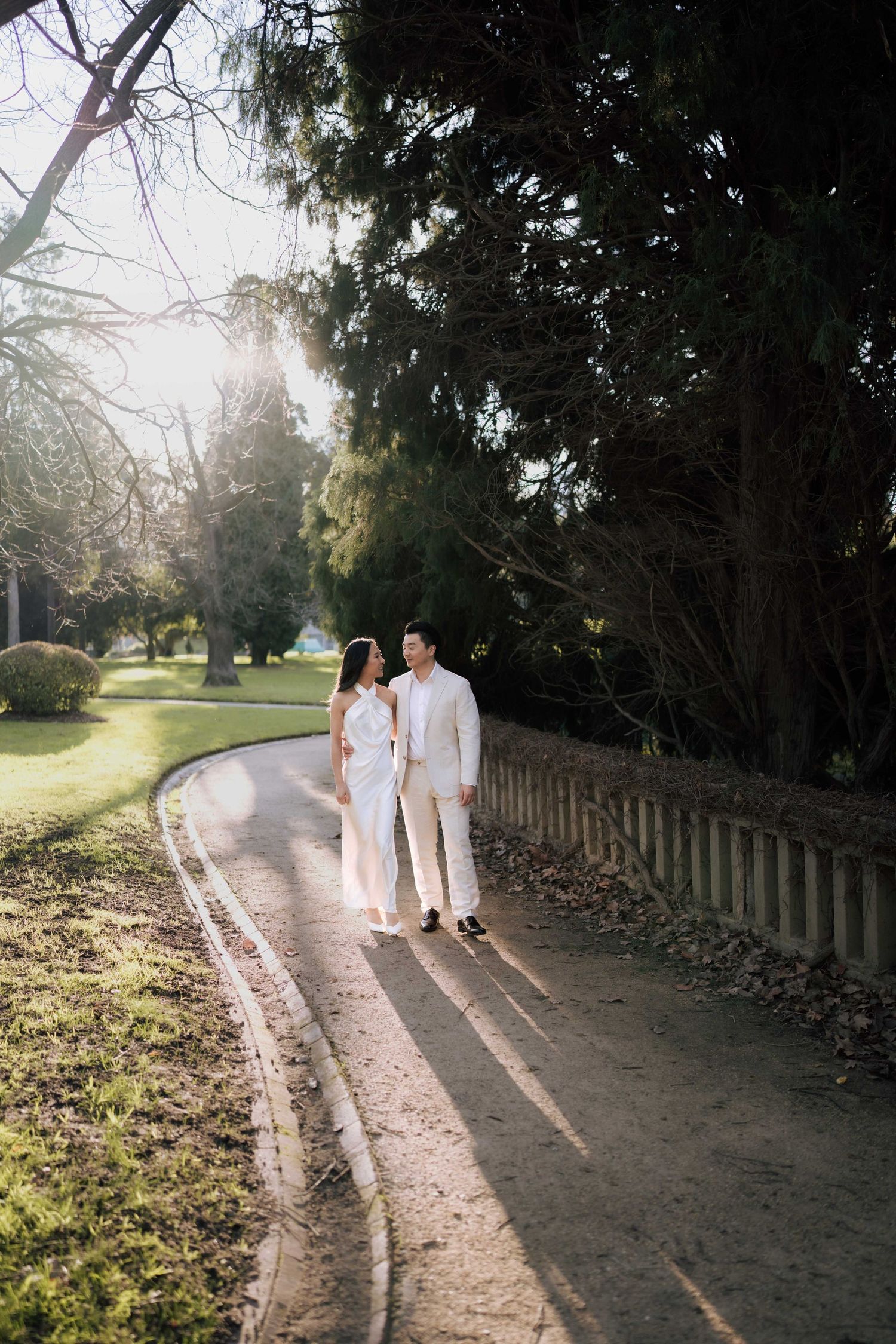 Couple in white outfits walking hand-in-hand along a sunlit park path lined with tall trees and a wooden fence.
