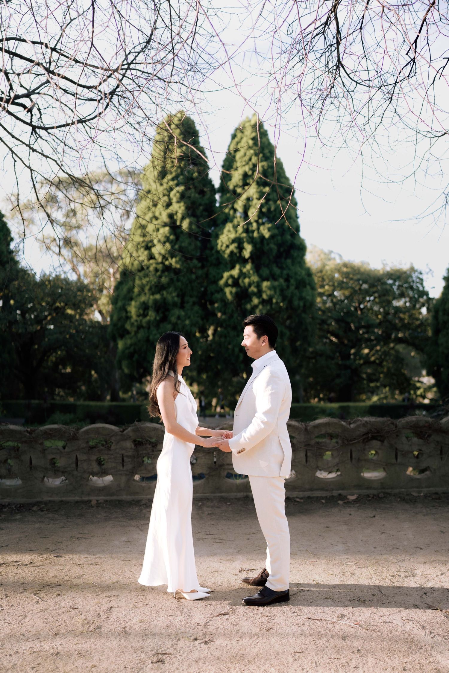 Couple in white attire holding hands in a garden with cypress trees and stone wall backdrop.