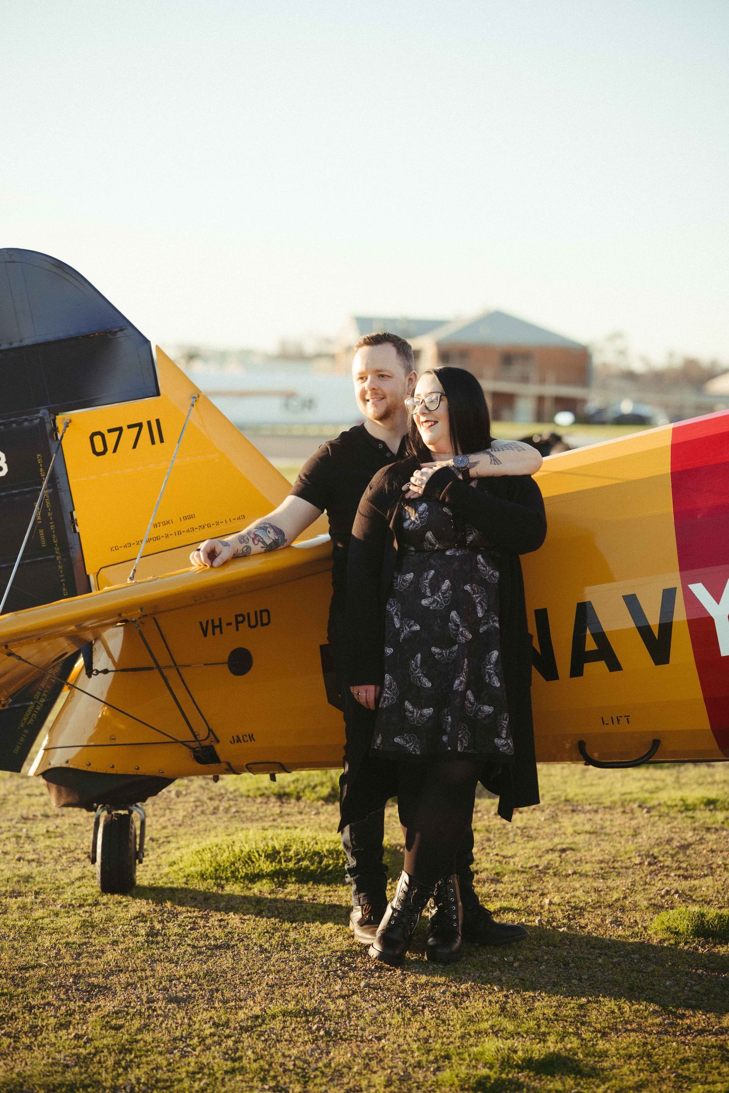 Couple smiling together beside a yellow Navy biplane on a grass airfield in warm sunlight.