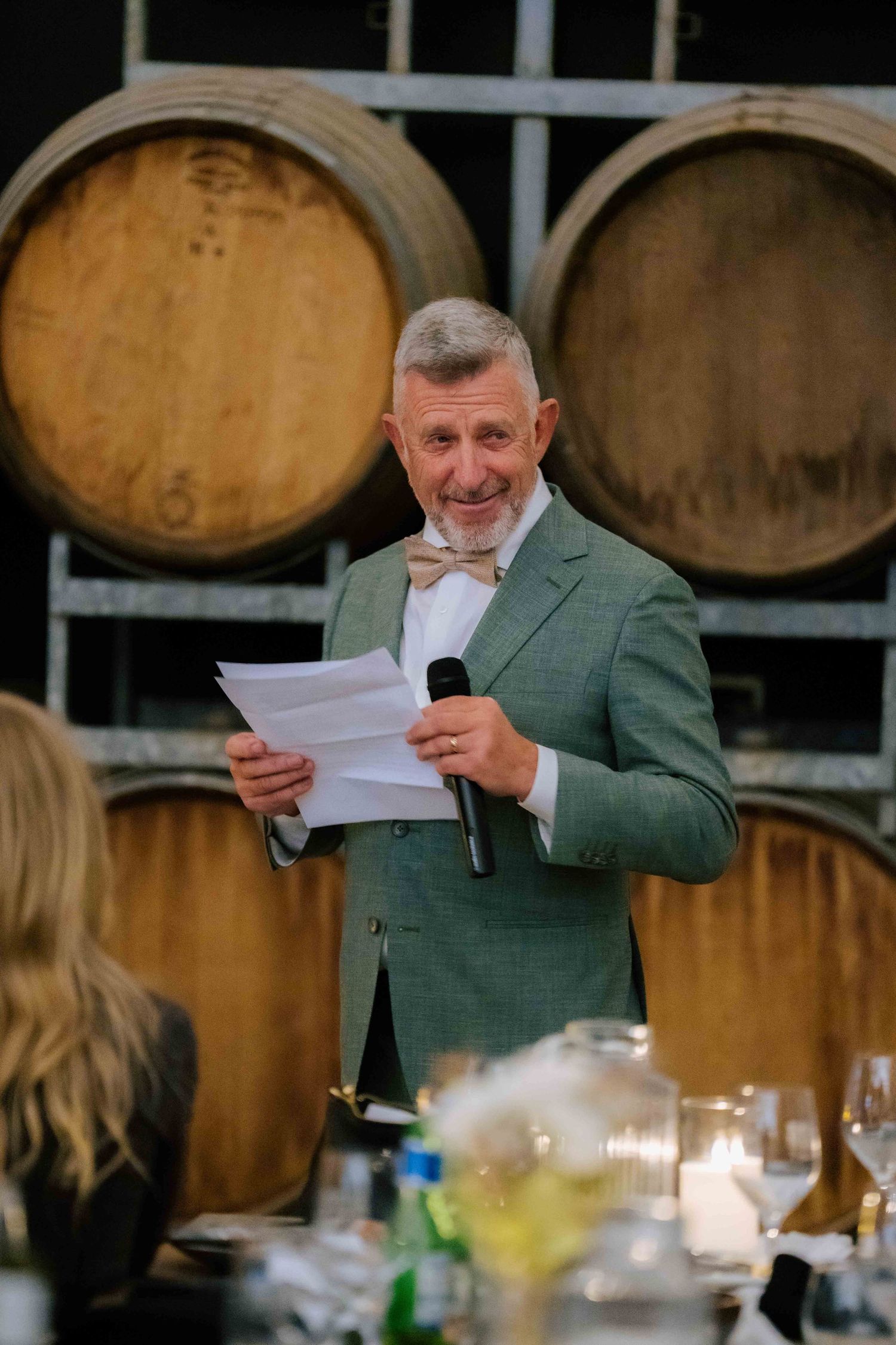 Man in green suit giving a wedding speech holding microphone near oak barrels.