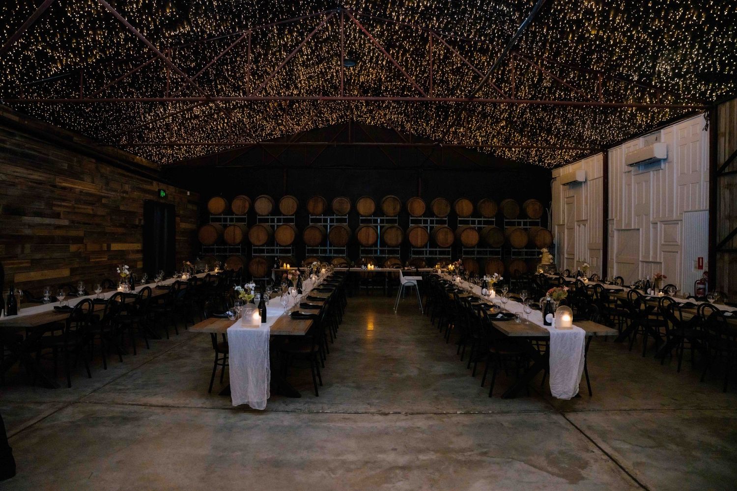 Stunning winery wedding reception hall with string lights on ceiling, long banquet tables, and wine barrel decor.