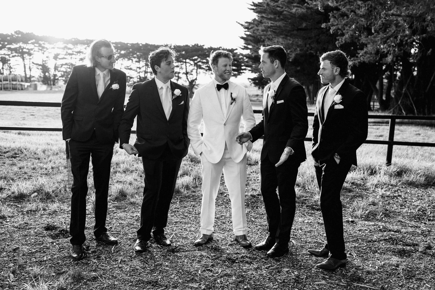 Black and white photo of groom in white suit with four groomsmen in dark suits outdoors.