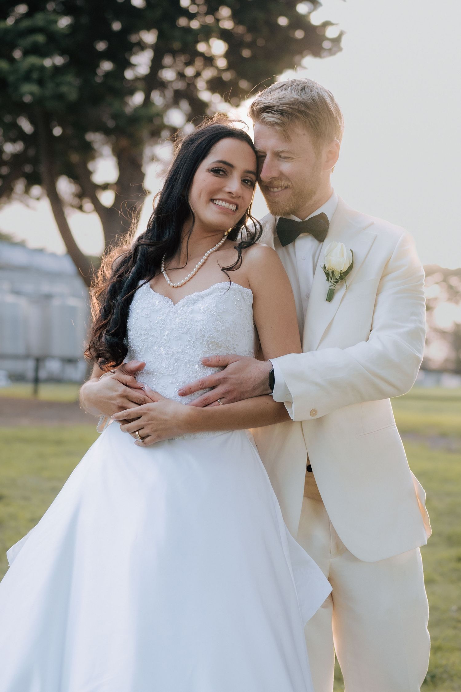 Bride in white strapless gown embraces groom in cream suit with bow tie during golden hour wedding portraits outdoors.