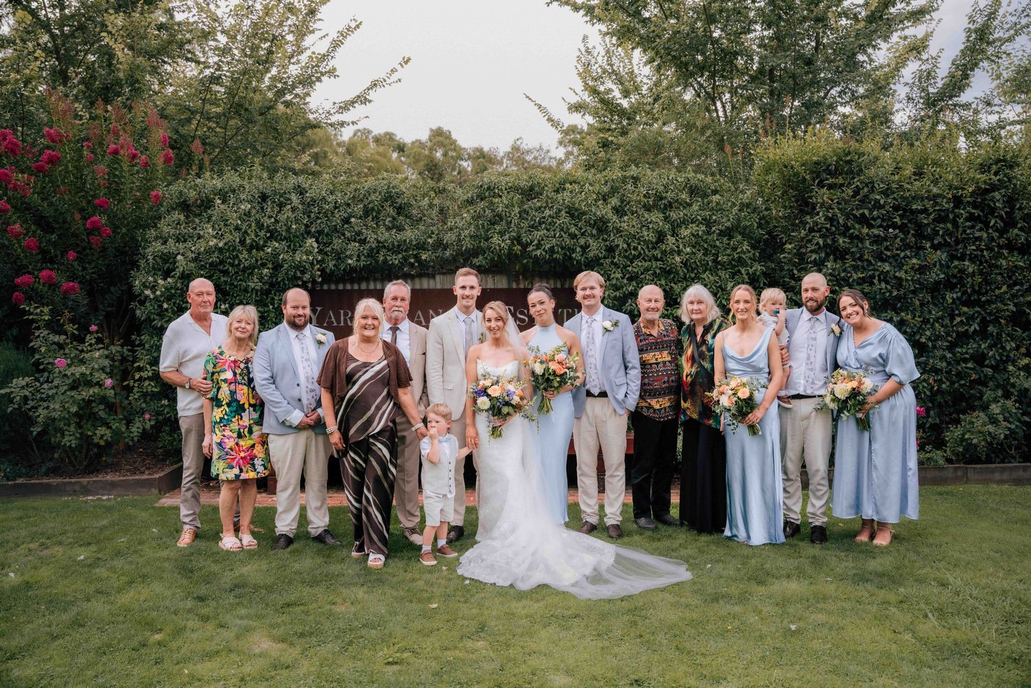 Wedding family portrait on lawn with bride and groom surrounded by relatives in front of lush green hedgerow.