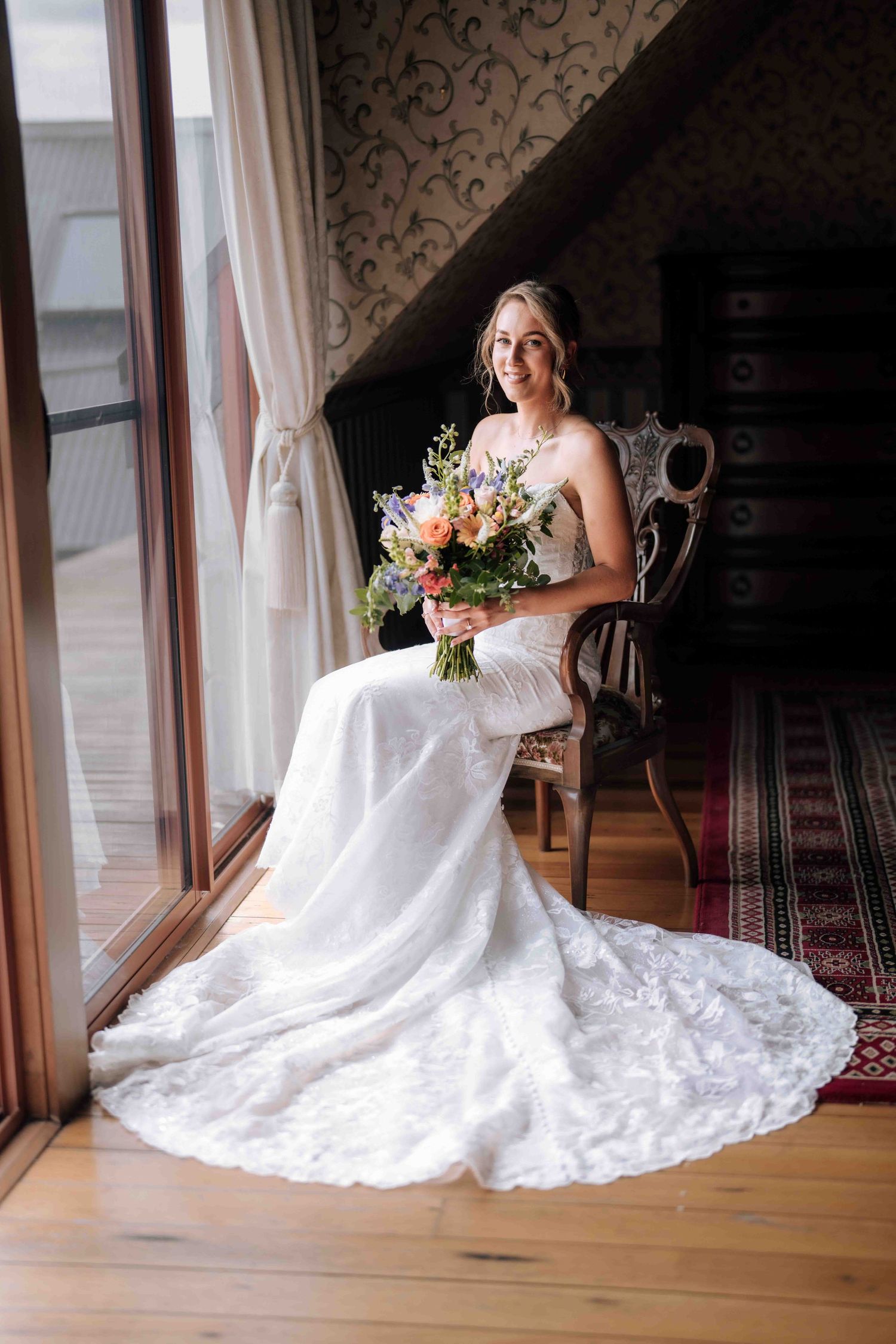 A bride in a white lace gown with a long train sits in an antique chair holding a colorful bouquet by a window.