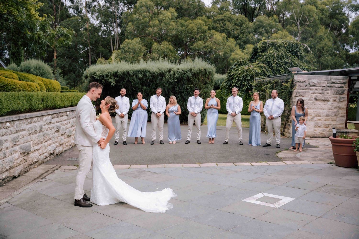 Bride and groom share romantic first kiss during first look on stone courtyard with wedding party.